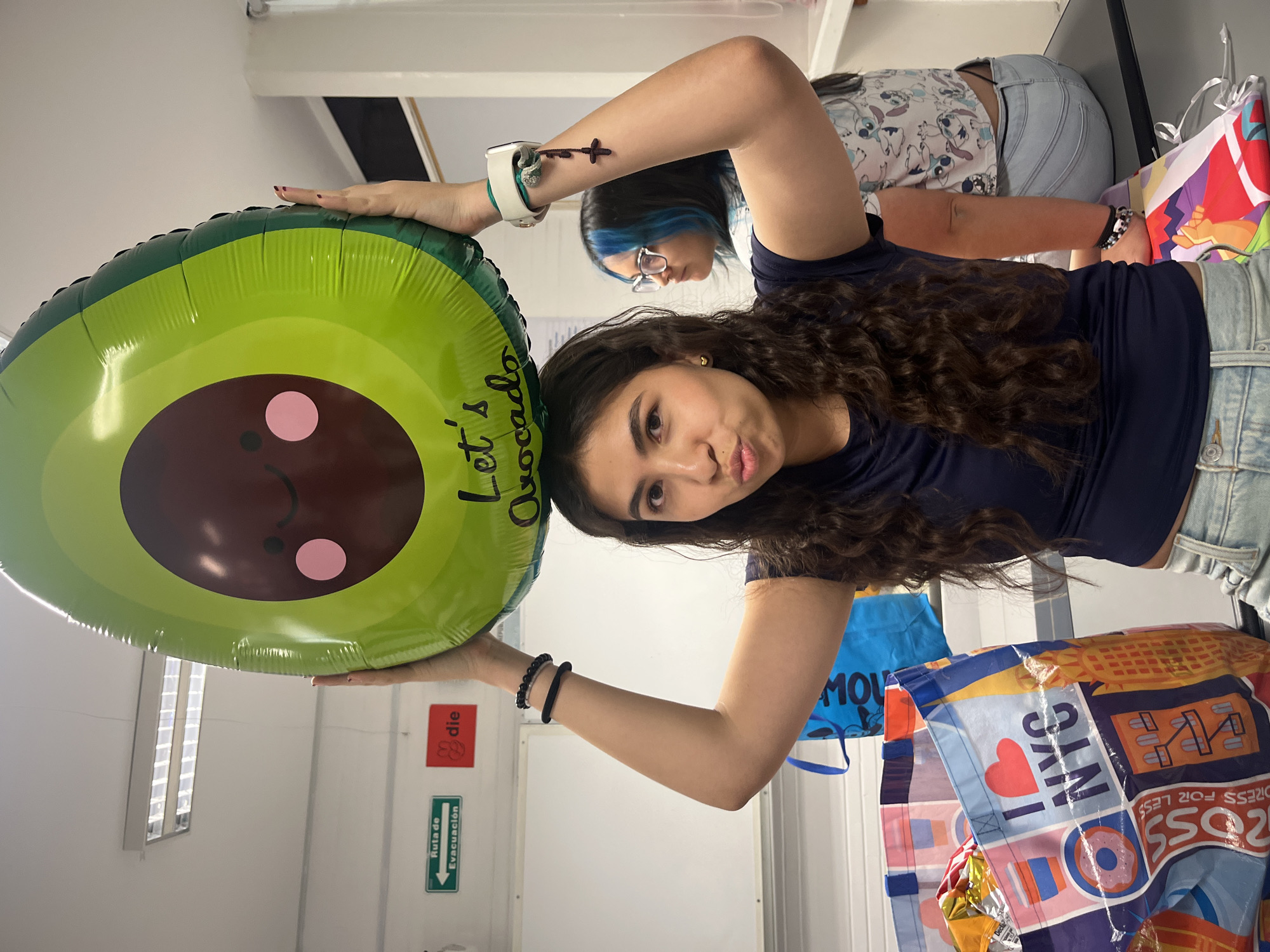 A woman with long, dark, curly hair is holding a large, round, avocado-shaped balloon with a brown pit that has a cute, cartoon face with pink cheeks. The balloon reads "Let's Avocado." The woman is wearing a dark blue shirt and light blue jean shorts. She is making a kissing face towards the camera. Behind her is a second woman with blue-streaked hair and glasses. There are various shopping bags nearby. One bag is blue with cartoon characters, and another has "I Heart NYC" printed on it with a "ROSS Dress for Less" logo. There is also an emergency exit sign visible in the background. The location in the city of Unknown, Unknown, cannot be determined from the image.
