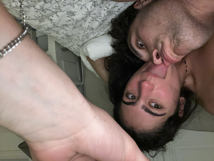 A couple lies intertwined in bed, their faces close and tongues touching in an intimate moment. The woman gazes at the camera, her dark hair contrasting with the floral pillow beneath her head. The man's face is partially obscured, his eyes focused on her. An arm with a bracelet extends into the frame. They're lying on patterned sheets, adding to the intimate, candid atmosphere.