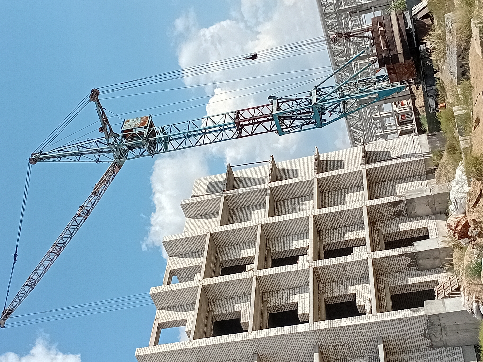 A construction site in Semey, Kazakhstan, features an unfinished, multi-story building constructed with white bricks and concrete balconies. A tall, blue tower crane with a red boom is positioned to the right of the building, its long boom extended over the structure. The crane has a small, enclosed operator's cabin and multiple cables. In the foreground, the ground is uneven, with piles of dirt, sandbags, concrete blocks, and a small wooden ladder. The sky above is blue with scattered white clouds. In the background, the skeletal framework of another structure is visible.
