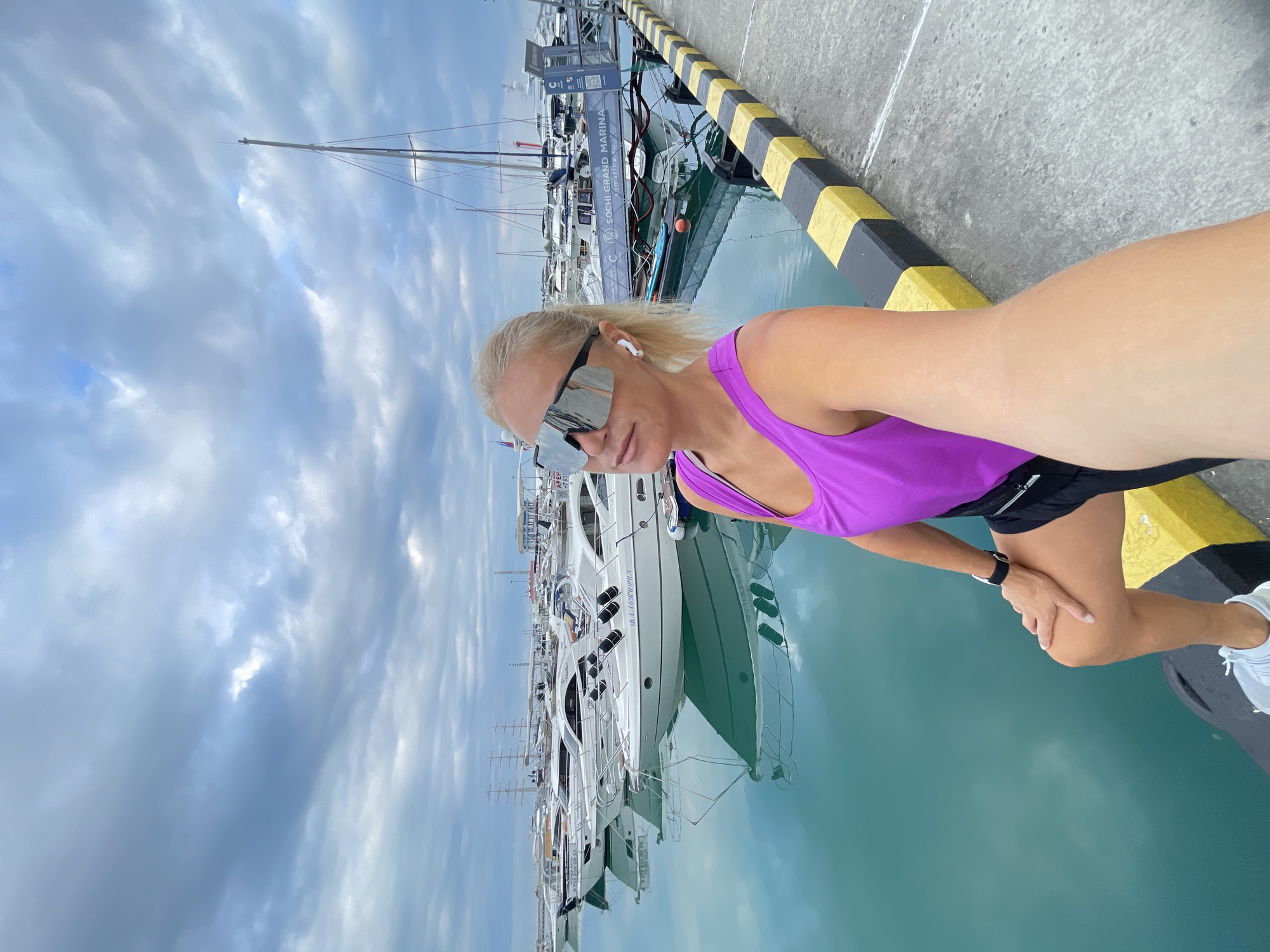 An athletic Caucasian woman with blonde hair, wearing a purple tank top, black shorts, large silver sunglasses, and white wireless earbuds, is taking a selfie in a marina in Sochi, Russia. She is seated on a yellow and black striped barrier, with her right hand resting on her right knee. Her left arm is extended forward, holding a phone, indicating she is taking the photo.
The background features a calm, turquoise body of water filled with numerous white yachts of various sizes. The sky above is overcast with dramatic, gray clouds. A sign on a gangway reads "SOCHI GRAND MARINA" with a website address. The overall impression is a scene of leisure and activity in a coastal city.