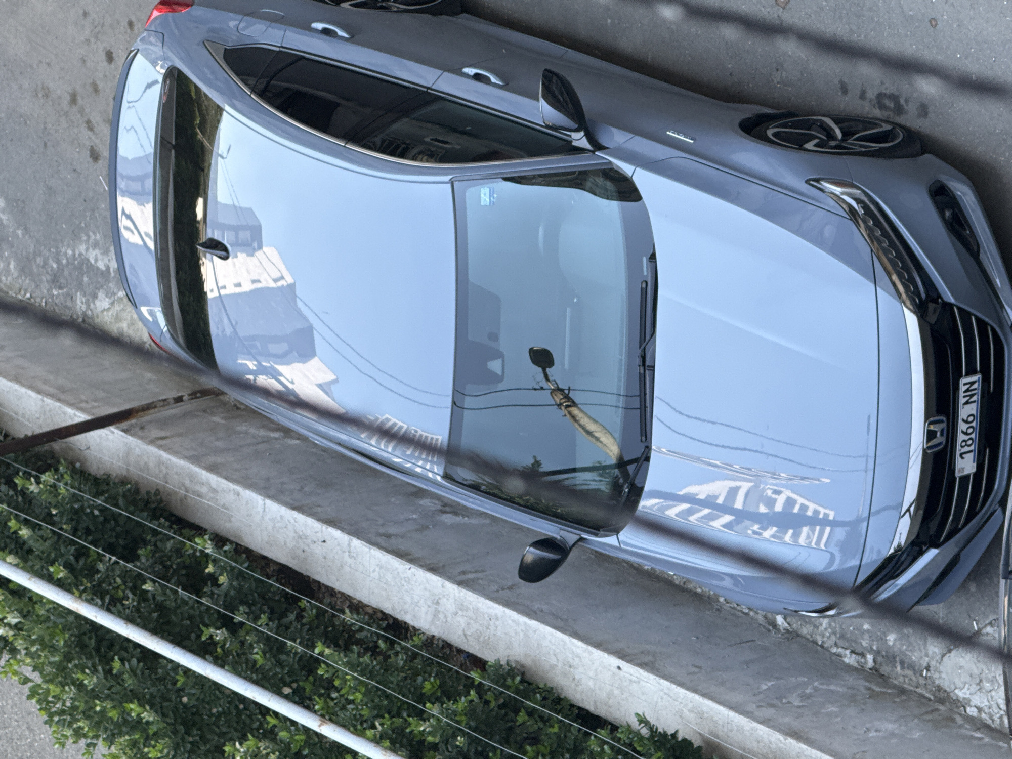 A grey Honda sedan is parked on a street in Tbilisi, Georgia. The car is viewed from a high angle, looking down at its roof and front. The car has a license plate that reads "1866 NN". The street is paved and there is a concrete barrier to the left of the car, with a bush growing behind it. Reflections of buildings are visible on the car's roof and windshield. A few dark wires or cables are visible in the foreground, obscuring parts of the image.