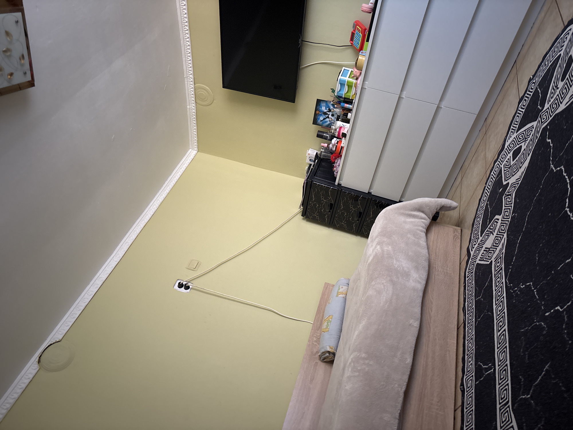 The image shows an interior space with a combination of domestic and potentially commercial elements. A television is mounted on the wall above a dark cabinet with storage, next to a light gray cabinet. An electrical cord runs from the wall outlet to the cabinet. Beside the wall is a piece of furniture, possibly a bed or couch, with a rolled item on top and a patterned black-and-white rug.
There is no information to place the location in Vetovo, Bulgaria.