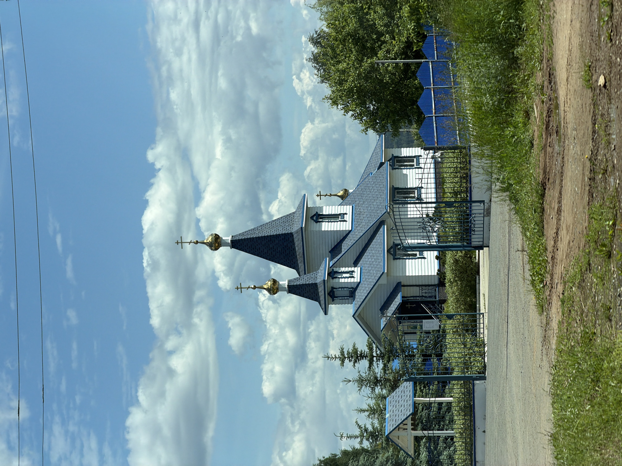 This is a daytime, eye-level view of a white and blue Eastern Orthodox church situated in Кварса, Russia, under a bright blue sky filled with numerous white cumulus clouds.
The church, constructed with white horizontal siding, features a dark blue shingled roof and prominent blue trim around its windows and architectural elements. It has two main spires, each topped with a conical dark blue roof, a small golden onion dome, and a golden Orthodox cross. A third, smaller golden onion dome with a cross is visible on a lower roof section towards the left. The church entrance, located centrally, includes a small porch with a blue roof and dark metal railings.
The church grounds are enclosed by an ornate dark blue or green metal fence. A matching, decorative metal gate stands open in the foreground, providing access to the property. To the left of the gate, within the fenced area, is a small, white A-frame structure with a blue shingled roof, resembling a shrine or canopy. Several young, conical coniferous trees are planted around the church, and mature green deciduous trees are visible on the right side, behind a corrugated blue metal fence.
The immediate foreground consists of a dirt or gravel road, showing patches of green grass and small puddles or damp areas. Two dark, thin power lines stretch horizontally across the top of the sky. No individuals are present in the scene.