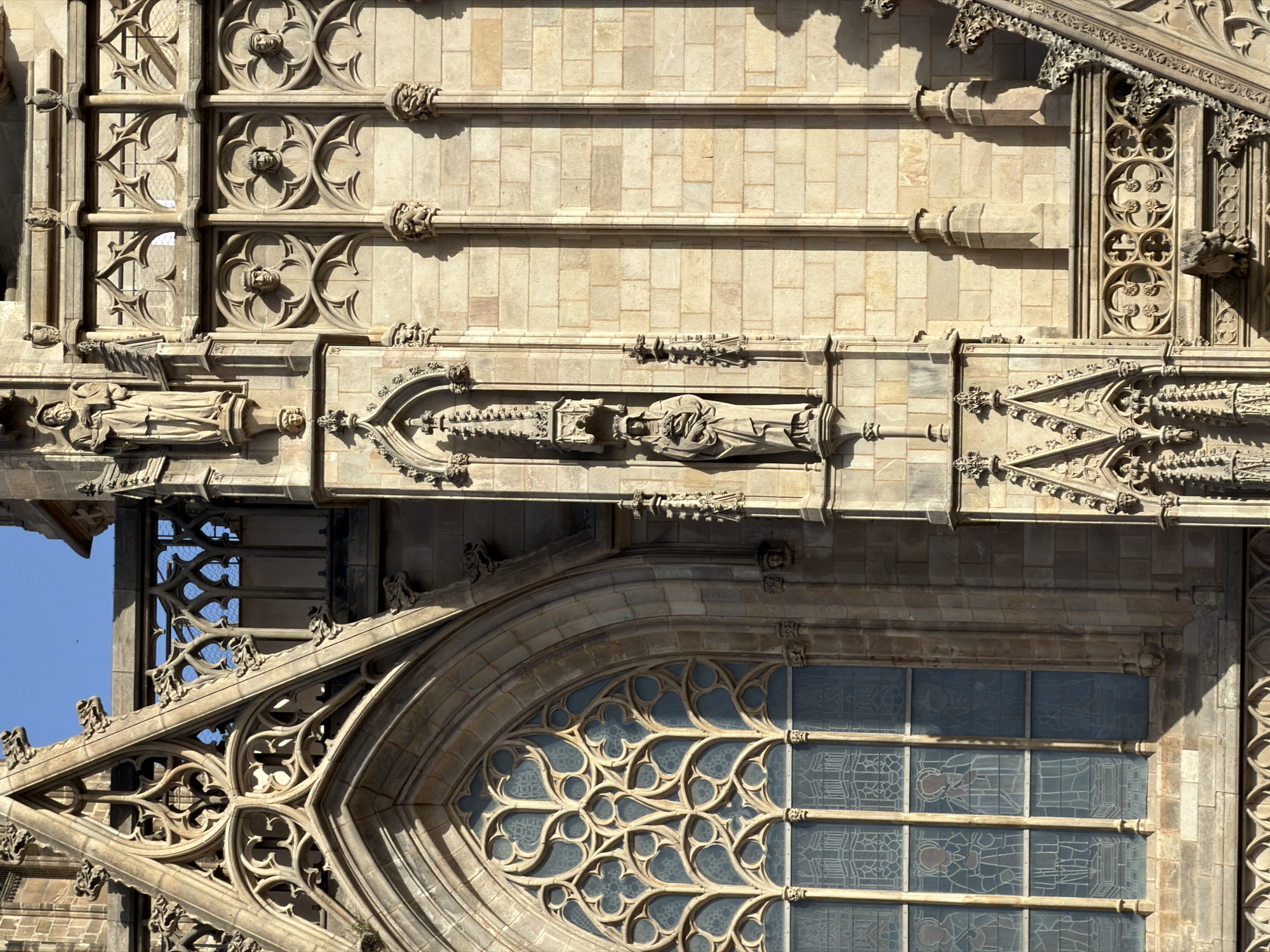 The image shows a close-up of the exterior facade of the Barcelona Cathedral in Barcelona, Spain. The architecture is Gothic, characterized by intricate stonework, pointed arches, and decorative tracery. The facade features several statues set within niches, including a figure holding a child. There are also decorative elements like gargoyles and carved faces integrated into the stonework. A large, ornate stained-glass window with geometric patterns is visible on the left side of the image. The stone blocks forming the wall are light brown and weathered, with shadows cast by the architectural features, indicating it is daytime.