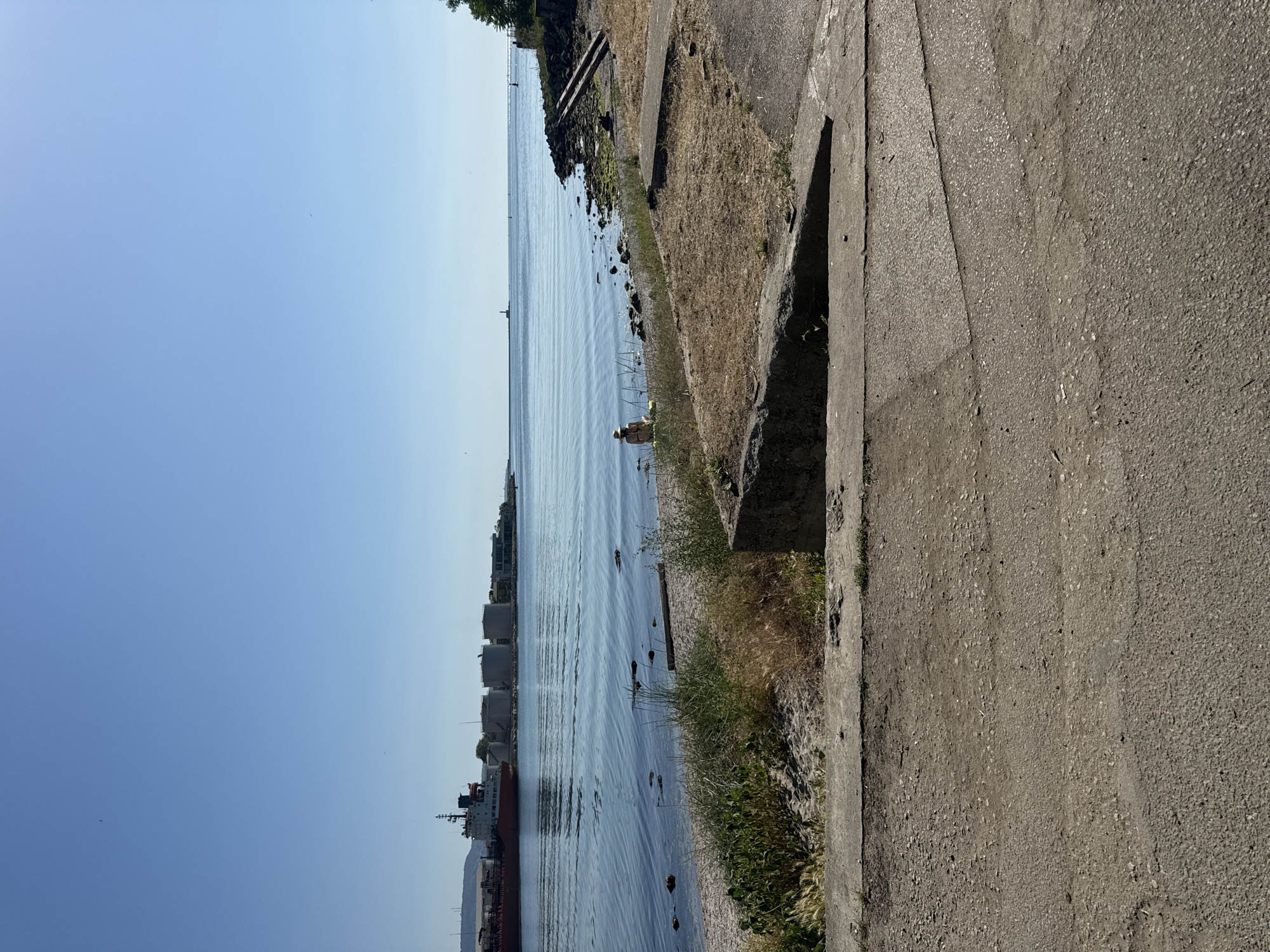 A bright, sunny day with a clear blue sky over a calm body of water. In the distance, a large red cargo ship is docked near industrial storage tanks. A person wearing a straw hat is sitting on a rocky beach, facing the water. The foreground consists of a concrete pathway leading down to the beach.