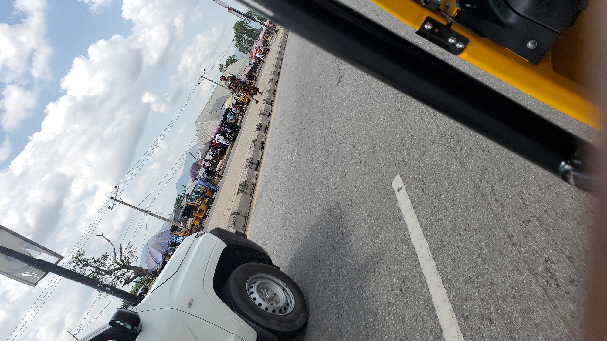 The image depicts a street scene in Jalingo, Nigeria. A white SUV is parked on the left side of the road. The road itself is paved and has a white lane marker. On the right side of the road, there is a sidewalk with various stalls and people. Several yellow tricycles, commonly known as Keke Napeps, are parked along the sidewalk, some appearing to be loading or unloading passengers. People are seen walking and standing, some under umbrellas. Power lines and utility poles are visible in the background, along with a large tree. The sky is partly cloudy. The foreground of the image is partially obscured by the black and yellow frame of what appears to be the interior of a vehicle, possibly another tricycle. The scene suggests a busy marketplace or transport hub.