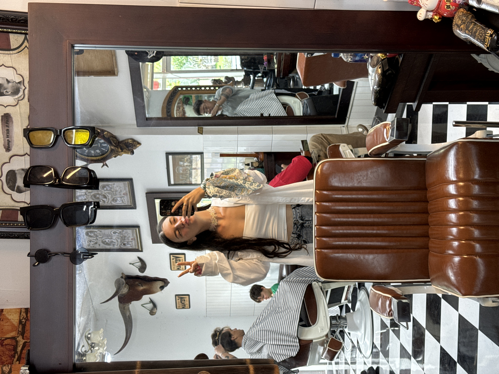 In a barber shop in Phan Thiết, Vietnam, a woman is taking a selfie in a mirror. She is wearing a white tube top, a white unbuttoned shirt, and denim jeans. She is holding a phone in her right hand and making a peace sign with her left hand. Her hair is long and dark. In the background, a barber is cutting a customer's hair. The barber shop has a vintage aesthetic, with a checkered floor, leather barber chairs, and various antique decorations, including a bull's head with horns mounted on the wall.