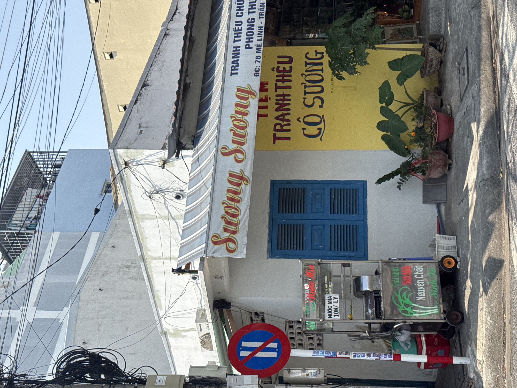 The image depicts a street scene with a business named "Sương Sương" located at 76 Mê Linh - Trần. The business appears to specialize in embroidery ("TRANH THÊU"). Adjacent to the building, a mobile vendor sells sugar cane juice ("NƯỚC MÍA") for 10,000 VND per glass. A "no parking" traffic sign is present near the vendor. Plants are placed in pots near the building. Utility lines and other buildings are visible in the background. The city is identified as Nha Trang based on signage.