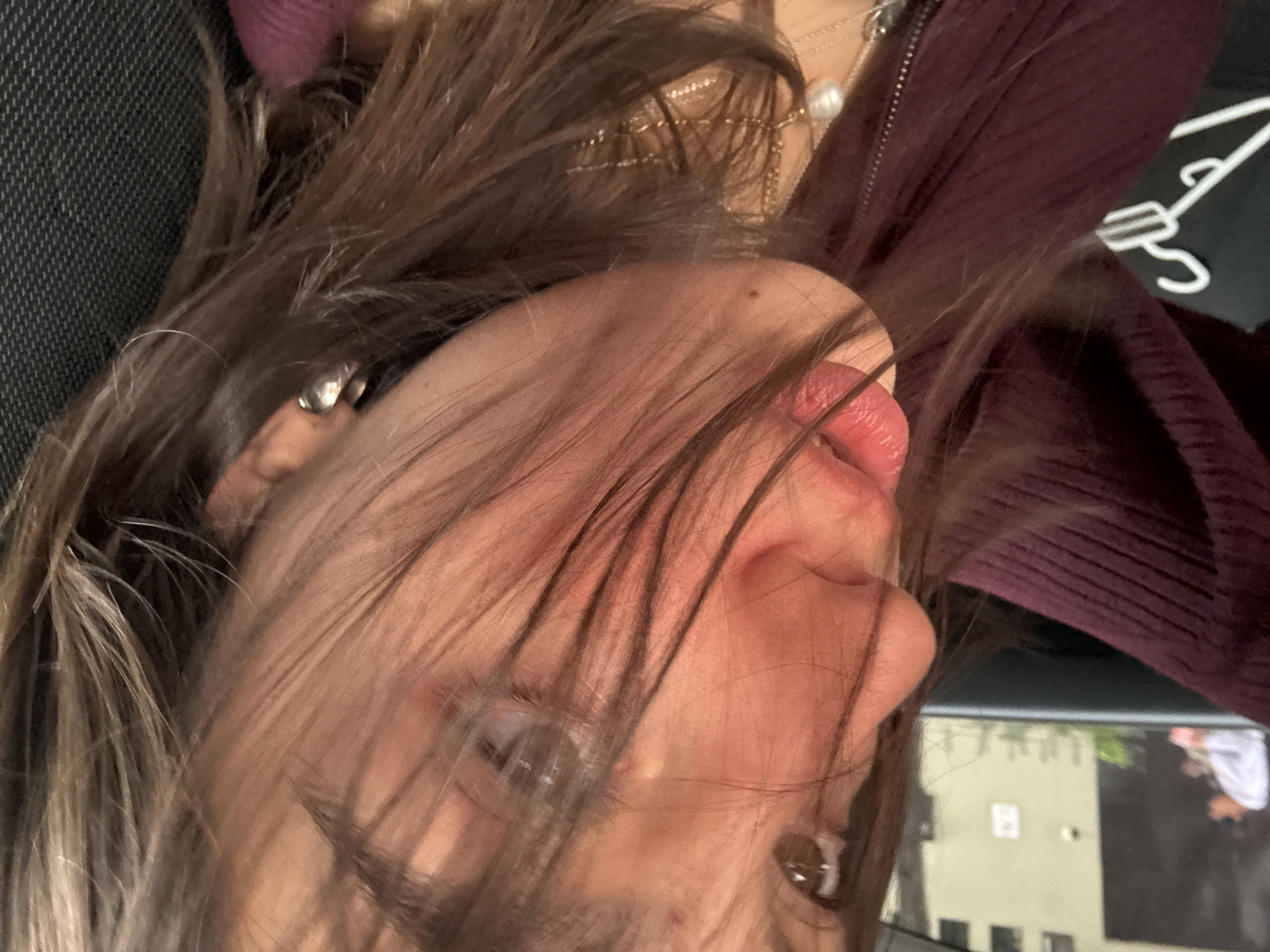 The image presents a close-up view of a person's face, partially obscured by strands of brown hair. The person appears to be wearing a dark burgundy colored zipper jacket and multiple necklaces. Part of the person's ear is visible, with what appears to be an earring. The background is blurry, revealing a cityscape that, based on the architecture style and color palette, is identifiable as Mexico City, Mexico.