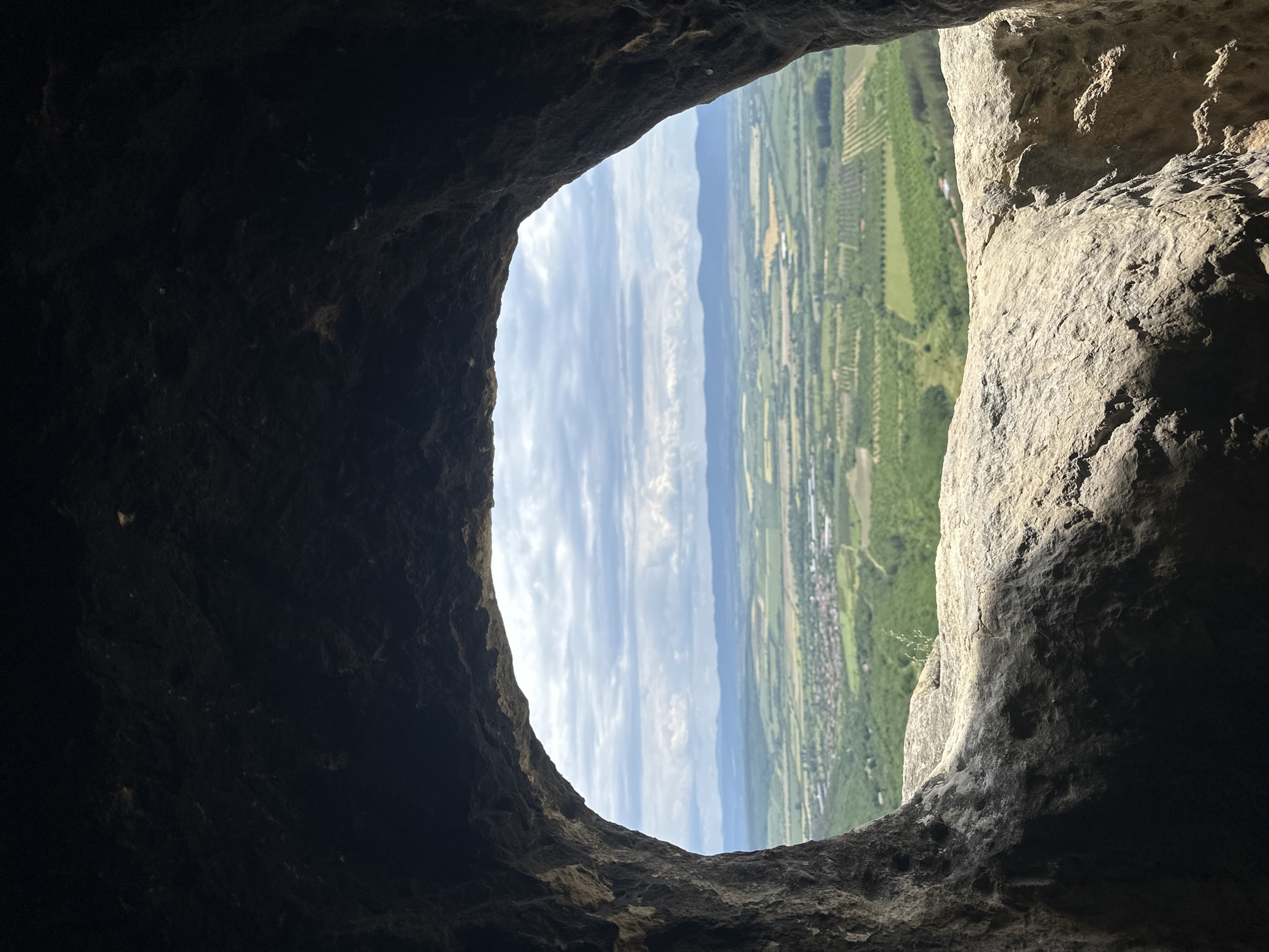 The image shows a view from inside a dark cave, looking out onto a landscape of fields, trees, and a distant town. The town is located in the Shumen Province of Bulgaria. The sky is visible above, with scattered clouds. The edges of the cave opening are rough and uneven.