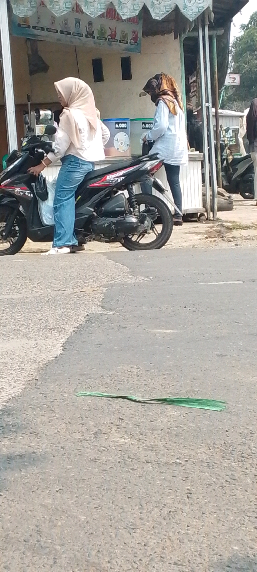 In a street scene in Cilaku, Indonesia, two individuals are near a small roadside stall. On the left, a person wearing a light pink hijab and a light pink long-sleeved shirt, along with blue jeans, is seated on a black motorcycle. Their right hand is near the motorcycle's handlebars. To the right, another person, partially obscured, wears a patterned brown and black headscarf, a light blue long-sleeved shirt, and dark pants. This individual appears to be interacting with the stall. Behind them, the stall displays several containers with "6.000" marked on them. The motorcycle is positioned on a paved surface adjacent to a dirt and gravel area. In the foreground, a green plastic strip lies on the asphalt. The background includes the stall structure and vegetation.