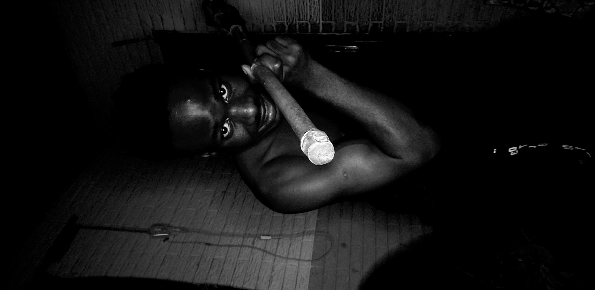 The image depicts a person lying on a wooden floor in a dimly lit room. The person, identified as male, has dark skin and is holding a metal pipe with both hands. The pipe is positioned diagonally across their chest. The background is dark, but a wall with some textured surface is visible behind the person's head. An electrical cord is visible on the floor near the bottom left corner. The location is specified as Narayi, Nigeria, but there are no visual cues to confirm this.