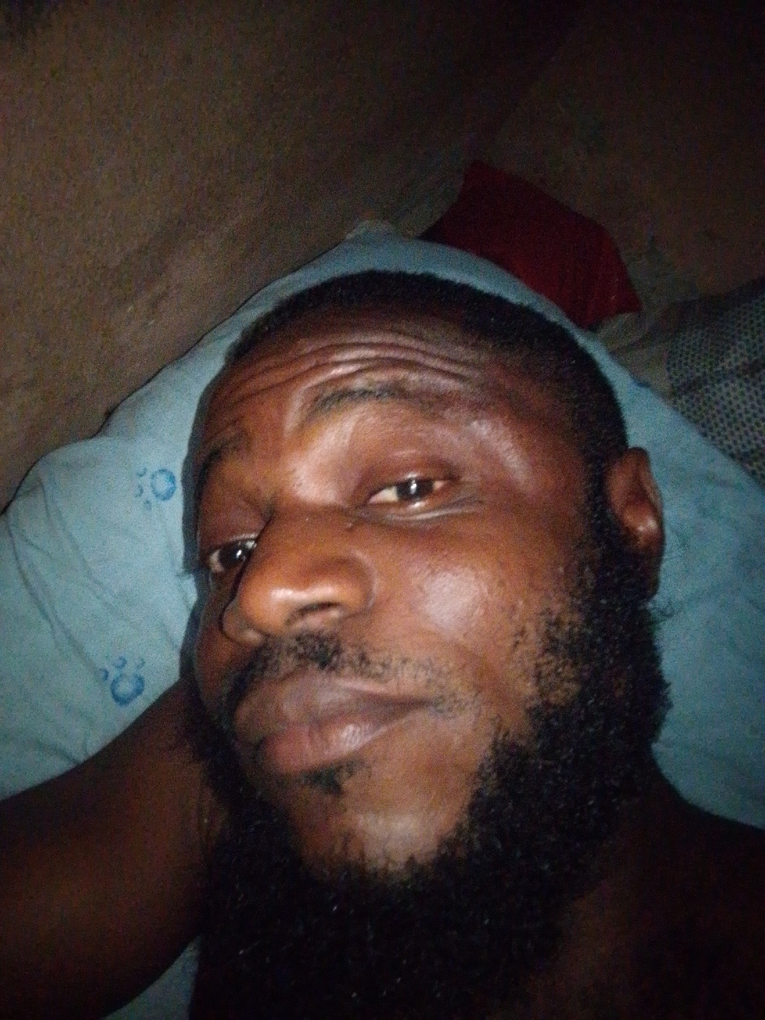 A close-up selfie of a dark-skinned man with a dark beard and short hair. The man is lying on a light blue pillow with a pattern, likely in bed, indoors. A red object is visible in the upper background. The lighting is dim. There is no evidence to confirm the location as Ajaokuta, Nigeria.