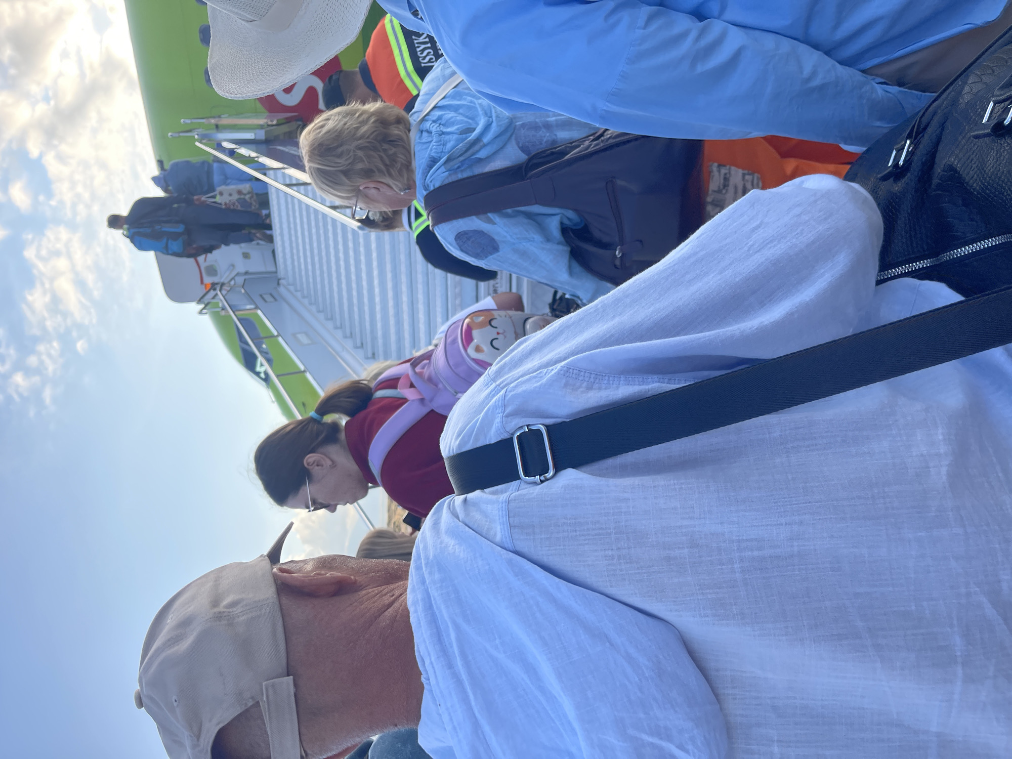 In Choktal, Kyrgyzstan, a group of people are boarding an airplane via a mobile staircase. The foreground features the back of a person wearing a light-colored cap and a white linen shirt, with a black strap from a bag visible across their shoulder. To their right, a woman with blonde hair, wearing glasses and a light blue hooded jacket, carries a dark brown shoulder bag. Behind her, other individuals are also ascending the stairs. The airplane is predominantly green, with some red branding visible. The sky above is bright and partly cloudy, suggesting it is daytime. The scene depicts a common travel moment at an airport.