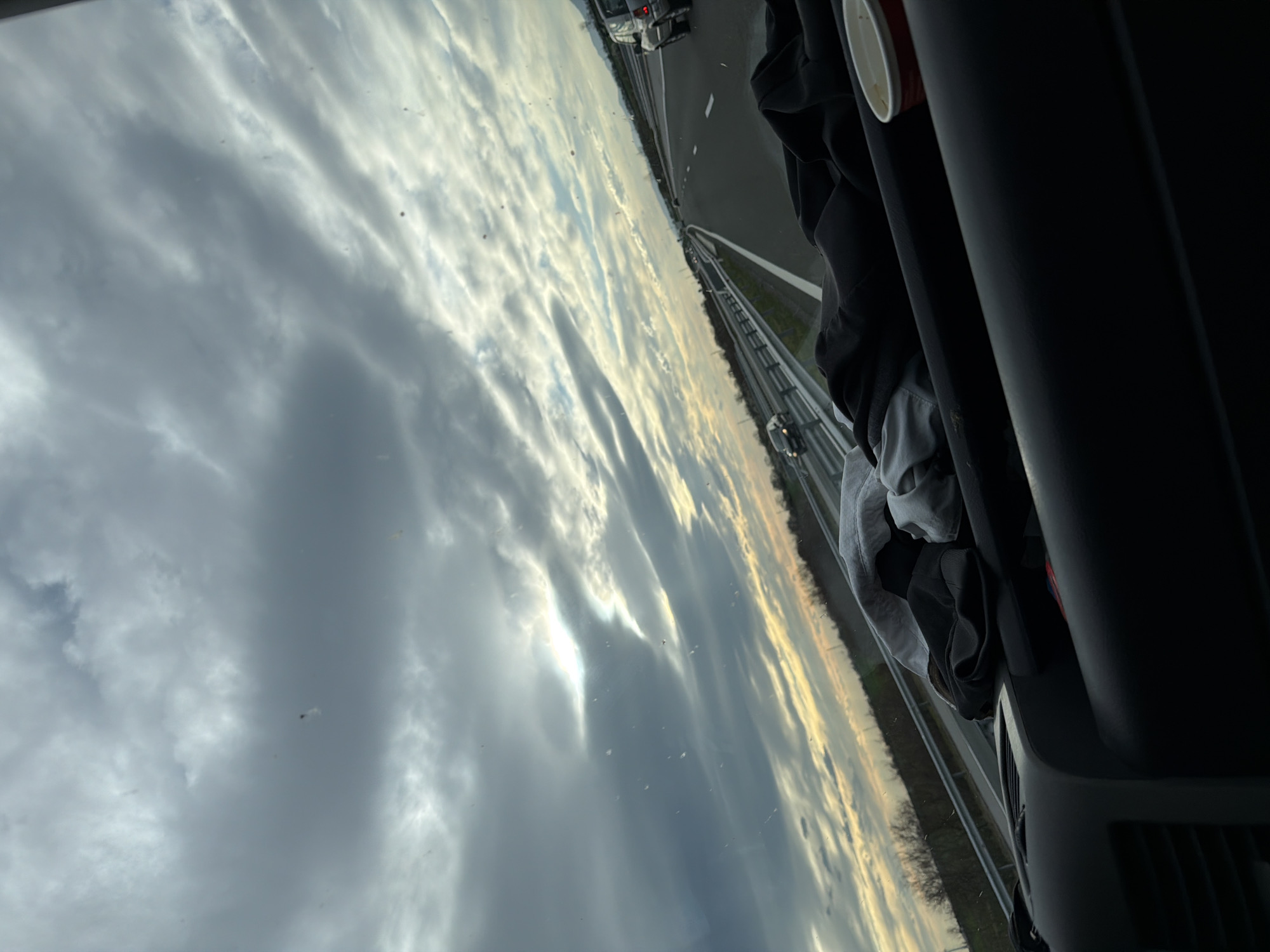 The photograph shows a view from inside a vehicle, likely a car, traveling on a multi-lane highway. The highway has a median barrier. Other vehicles are visible on the road. The sky, filled with numerous clouds, dominates the left portion of the frame and appears to be reflected in the vehicle's windshield. The interior of the car is partially visible on the right, with clothing and a cup placed near the dashboard. The image was not taken in Vodenichane, Bulgaria; the highway environment indicates a more developed infrastructure not typically found in smaller villages.