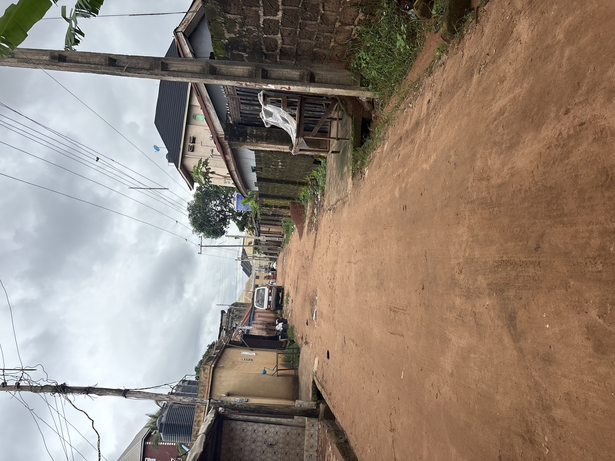 The image depicts a narrow, unpaved street in Nkpor, Nigeria, under a cloudy sky. The road is made of red earth, showing tire tracks from vehicles. On either side of the street are buildings constructed from concrete blocks and corrugated metal, with some showing signs of disrepair. On the left, a building features a patterned facade and a water tank on its roof. Further down the street, a white van is parked. On the right, a stone wall borders a property with a metal gate. A wooden table covered with a white plastic sheet stands near the gate. Vegetation, including tall grass and some trees, is visible along the sides of the street. The scene appears to be a residential or commercial area in a developing urban environment.