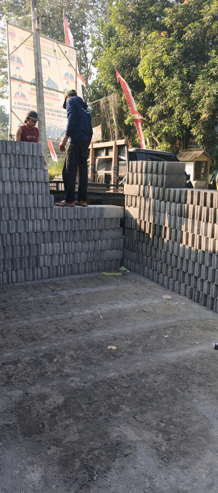 In Kasiyan, Indonesia, two individuals are present at a construction material site. The scene is characterized by stacks of gray concrete bricks, which are the primary objects. One person, wearing a dark blue jacket and black pants, stands on top of a stack of bricks. The other person, wearing a red tank top and a dark cap, is also standing among the bricks. In the background, a truck is visible, along with a small, light-colored building. Additionally, there are posters with images of people and text on a pole, along with Indonesian flags. The environment appears to be outdoors, with trees and foliage visible in the background.