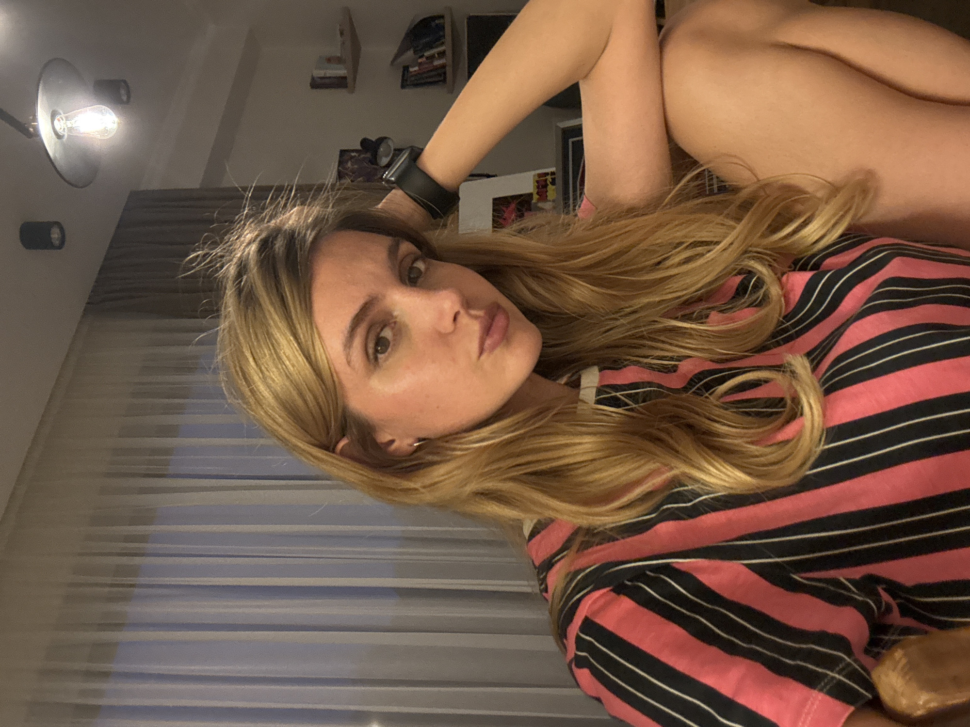 A young woman with long, wavy blonde hair is featured in this indoor portrait, looking directly at the camera. She is wearing a pink and black striped short-sleeved top. On her left wrist, she has a dark-colored smartwatch or wristband. Her pose suggests she is reclining or lounging, with her right arm raised above her head and parts of her leg or hip visible in the upper right.
The background shows an indoor setting, likely a room, with shelves holding various items, including what appear to be books. An illuminated light fixture with an exposed bulb is visible in the upper left, casting a glow. Other light sources or fixtures are also faintly discernible. The lighting is predominantly artificial, indicating the photo was likely taken in the evening or at night. Horizontal lines in the background might suggest blinds or a window. The overall composition is angled, creating a casual and intimate atmosphere. The woman's expression is neutral, with full lips. No visible text is present in the image.