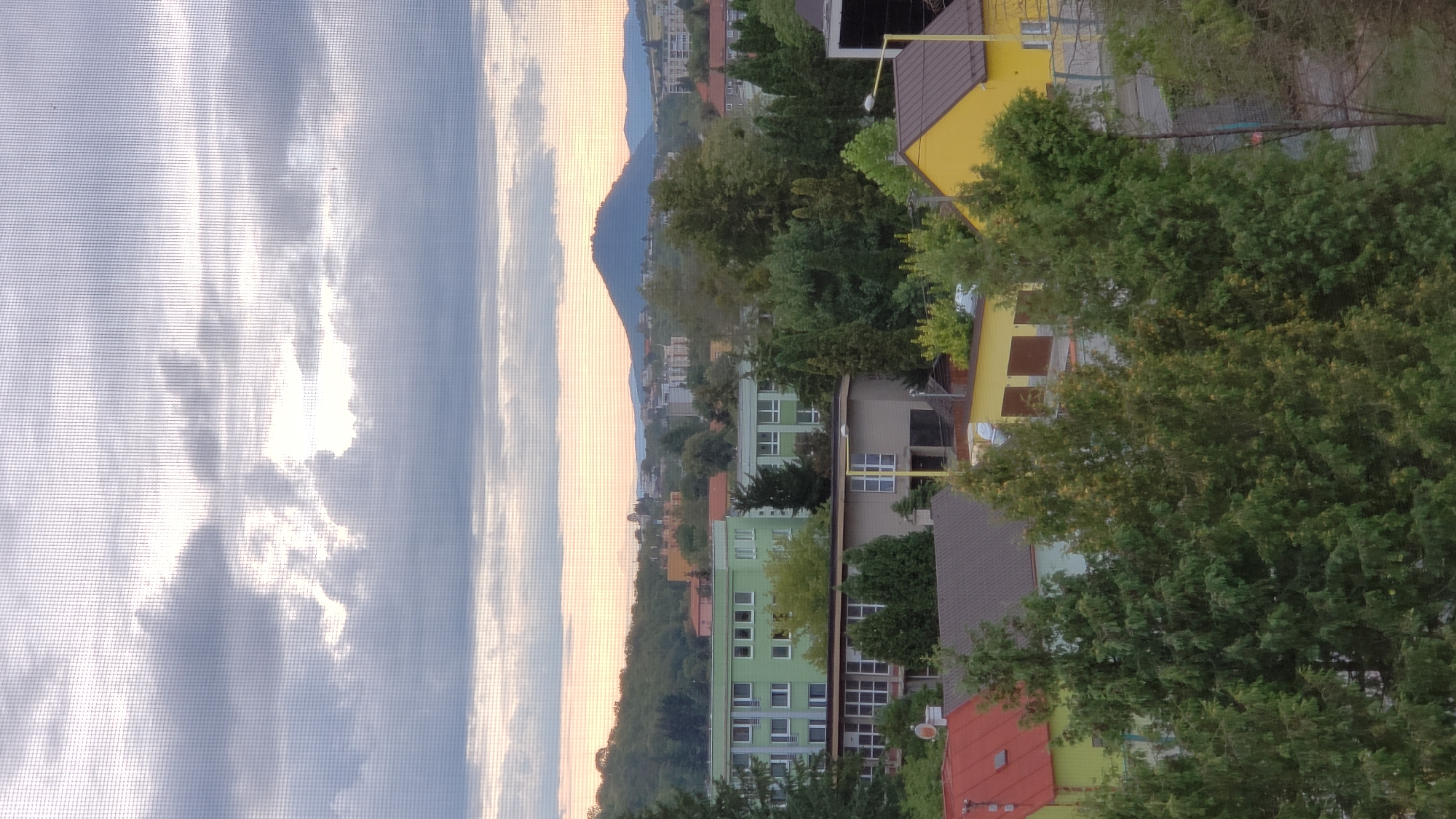 This image, taken from a window with a visible screen, depicts a view of a city, likely Prešov, Slovakia, during daylight hours. The scene is dominated by a mixture of buildings and dense foliage. In the foreground, several evergreen trees with dense, dark green foliage are prominent. Behind the trees, various buildings are visible.
The most noticeable structures include a long, light green building with multiple windows arranged in horizontal rows, and another similar, but shorter, green building. To the right of these, a mustard-yellow building with a brown roof and two large garage doors stands out. A smaller building with a red roof is visible to the left, partially obscured by trees. In the middle ground, a low-rise, brownish building with large windows and a visible yellow pole structure is present.
In the background, a hill or mountain with a rounded peak is silhouetted against a cloudy sky. The sky itself is overcast with a mix of light and dark gray clouds, suggesting potential for rain or a generally subdued atmosphere. The horizon line shows a range of buildings and trees, indicating a moderately sized urban or suburban area. The lighting suggests it is either early morning or late afternoon, with diffused sunlight. There are no people or vehicles clearly visible in the frame.