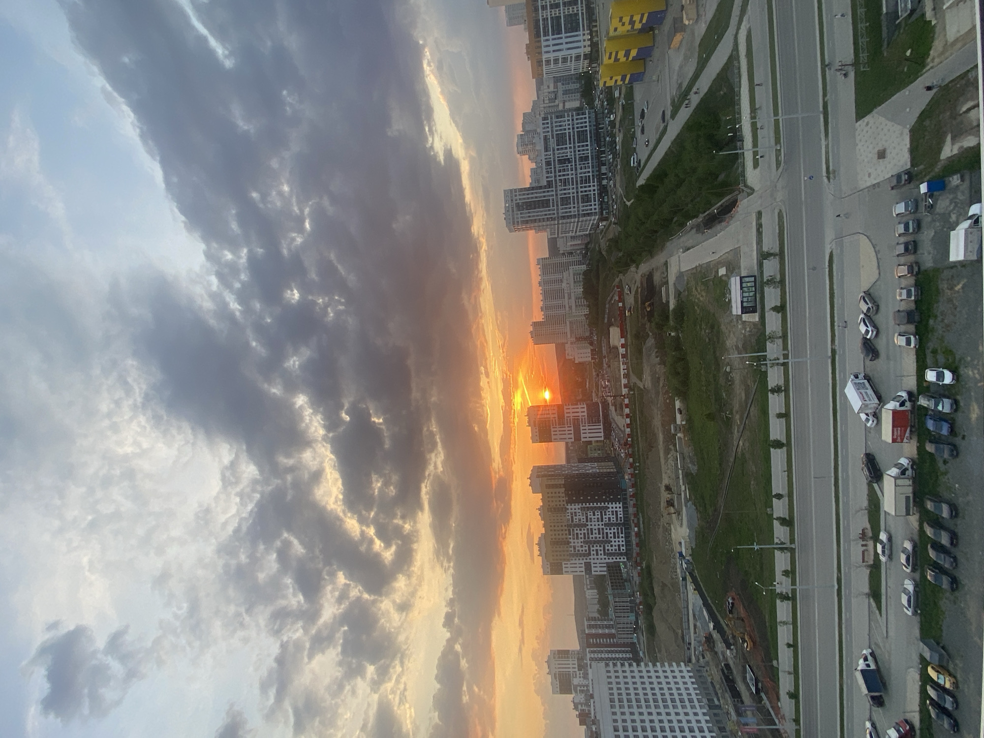 The image shows a sunset view of the Akademicheskiy district in Yekaterinburg, Russia, from a high vantage point. The sky is filled with clouds illuminated by the setting sun. Below the clouds, there are high-rise apartment buildings. There is a road running adjacent to the buildings. Parked cars line the street. A construction site is visible between some buildings.