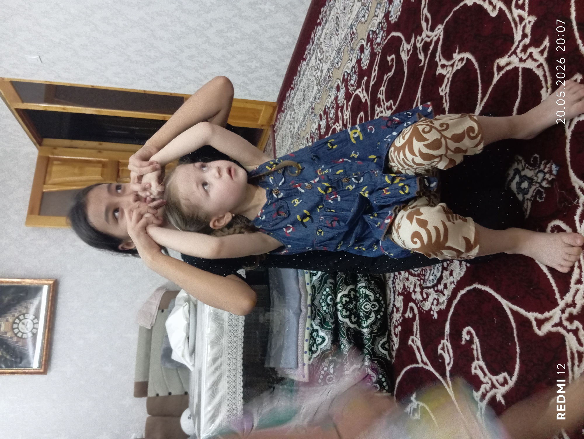 The image shows a young girl lying on her back on what appears to be a patterned carpet, with an older girl/woman leaning over her. The younger girl wears a blue dress and tan patterned leggings. The older girl/woman wears a dark-colored top. They are interacting, as the older girl/woman seems to be holding the younger girl's hands near her face. Behind them is a covered object that appears to be a bed or furniture. A picture frame is visible on the wall. The bottom right corner has "20.05.2026 20:07" and "REDMI 12" printed on it. The location is likely in Bekabad, Uzbekistan, based on the cultural elements of the image.