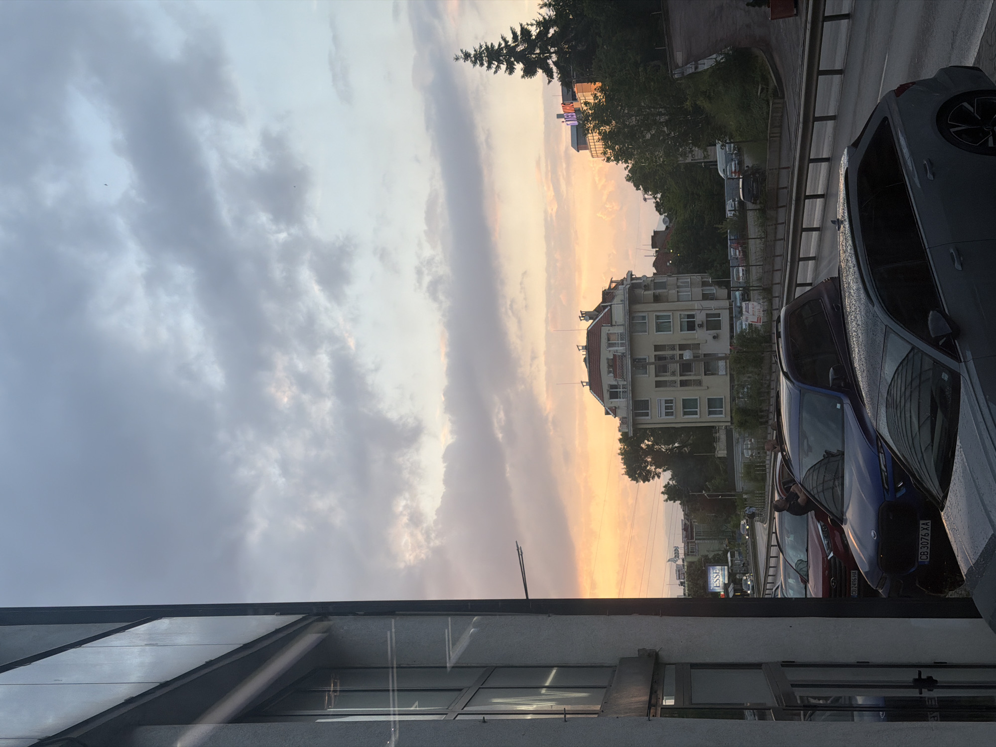 The image depicts a street scene in Sofia, Bulgaria, at dusk. In the foreground, the corner of a building with a glass façade is visible. A row of parked cars lines the street, including a gray car, a blue car with license plate "CB 3076 XA", and a red car with license plate "CB 7496 HA". The cars appear wet, suggesting recent rain. Across the street, a multi-story apartment building stands amid trees. Beyond, a building with "MEGAZONE" in lit-up letters can be seen in the background. The sky is filled with a mix of gray clouds and a gradient of orange and yellow light, indicating a sunset. The scene is lit by the natural light of the setting sun.