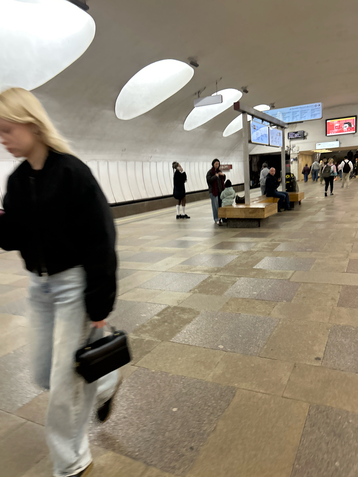 The image depicts a public space, likely a metro station in Moscow, Russia, characterized by its distinctive architecture. The ceiling features large, illuminated oval-shaped structures, creating a modern and somewhat abstract aesthetic. The floor is paved with large, rectangular tiles of varying shades of beige, brown, and grey, arranged in a grid pattern.
In the foreground, a young woman with blonde hair, wearing a black bomber jacket and light-colored wide-leg jeans, is captured in motion, walking from left to right. She is carrying a small black handbag in her right hand. Her face is blurred due to the motion blur of the image.
Further down the platform, several other individuals are present. A person in a dark outfit stands near a white wall. Another person, wearing a burgundy scarf and dark clothing, is standing next to a young child dressed in a light blue jacket. A man in a dark jacket and blue jeans is seated on a wooden bench, looking towards his left and holding a mobile phone. To his right, another wooden bench is occupied by several people, some standing and some sitting, appearing to be waiting. In the background, more individuals are seen walking along the platform, and several signs are mounted on a metal structure, including digital information displays and a rectangular screen showing a graphic. The overall atmosphere suggests a typical scene in a busy urban transit hub.