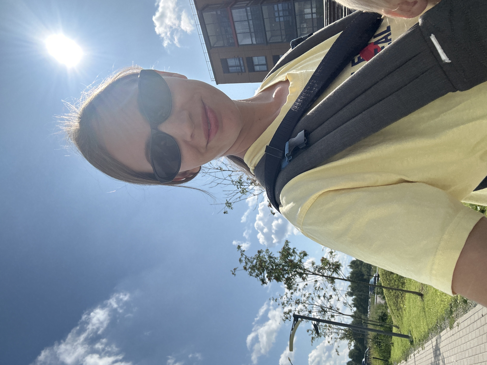 A medium-shot selfie shows a woman with brown hair, wearing sunglasses and a yellow t-shirt, with a baby carrier on her chest. She is outdoors on a sunny day, with a bright sun in the upper right corner of the frame. In the background, there is a modern apartment building, trees, and a sidewalk. The woman is smiling. The scene appears to be in Vsevolozhsk, Russia, given the context provided.