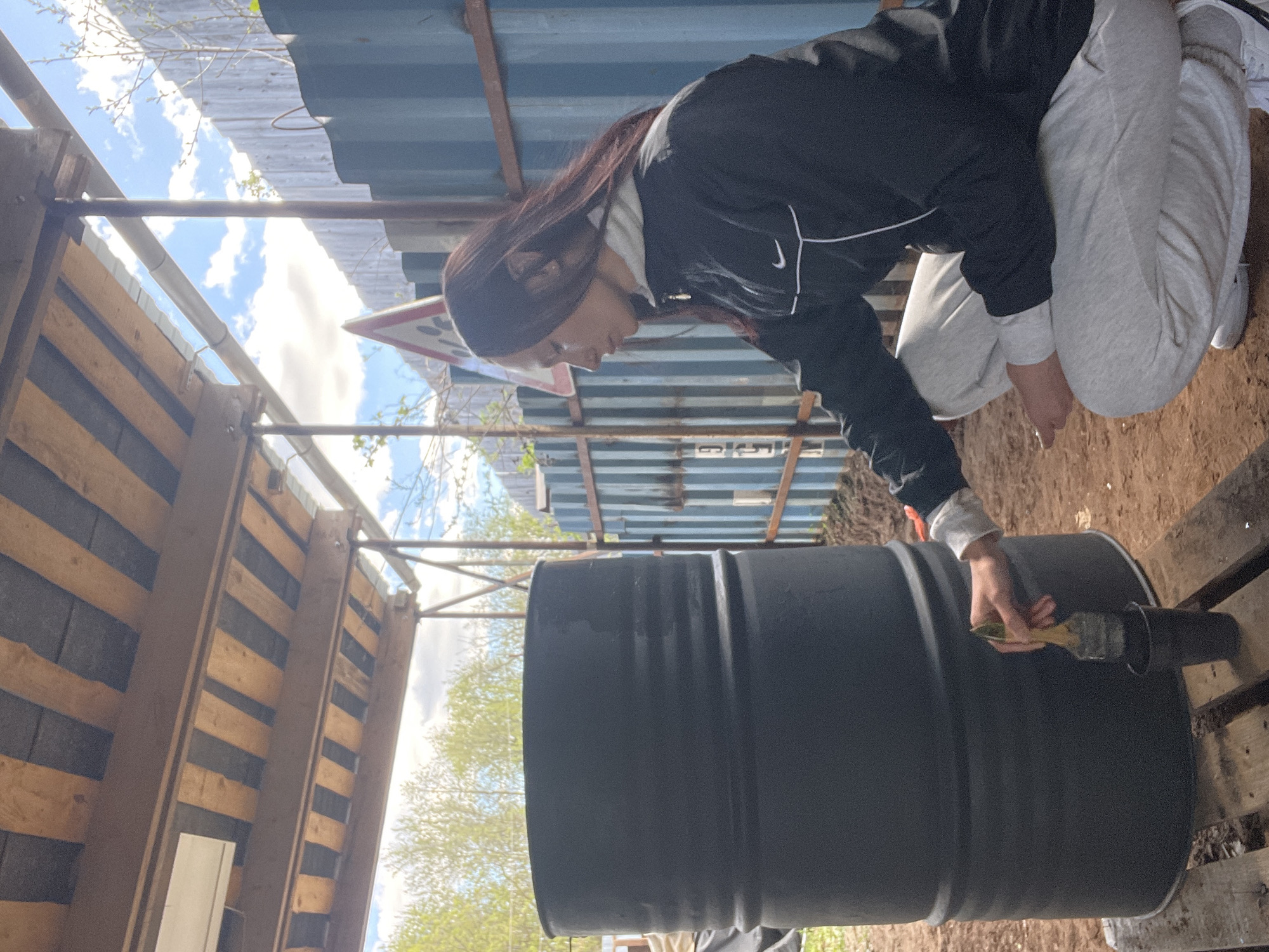 A woman in a black jacket and gray sweatpants is crouched down and appears to be painting a large black barrel with a brush. The barrel is resting on a wooden pallet. The surrounding environment includes what appears to be a corrugated metal roof structure and wooden walls. The sky with clouds is visible through gaps in the structures. The scene takes place outdoors, likely in Pervomaysky, Russia.