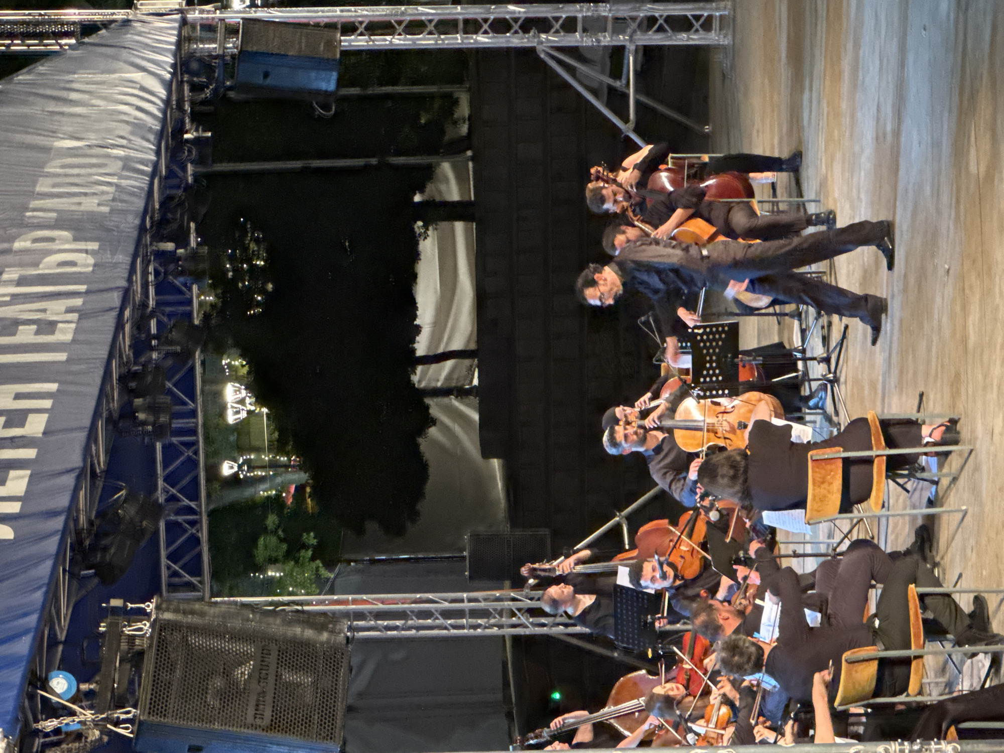 The image depicts an outdoor orchestral performance at the "Archaic Theater Apollonia" in Sozopol, Bulgaria. The orchestra members, dressed in black attire, are seated and playing various string instruments, including cellos and double basses, on a concrete stage. A conductor, also in black, stands on the right side of the stage, facing the orchestra and appearing to lead them.
In the foreground, several musicians are actively playing their instruments. A woman in the lower left is partially visible, seated and playing a cello. To her right, a man is also seated and playing a cello. Further back, several other musicians are playing cellos and double basses.
The stage is set up under a large blue canopy or tent structure that displays text in white letters. Several large speakers are suspended from the structure, and lighting equipment is also visible. Behind the orchestra, trees and illuminated streetlights create a backdrop. The overall atmosphere suggests a night-time event.