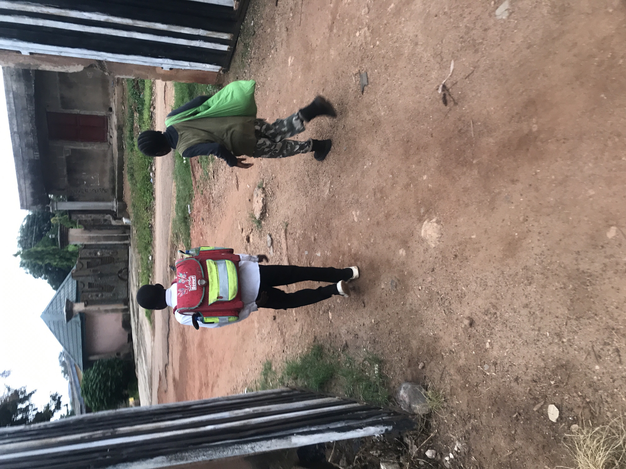 Two young boys walk away from the camera on a dirt path in Kabong, Nigeria. The boy on the left, wearing a white shirt and black pants, carries a red and yellow backpack. The boy on the right, in a green vest over a dark shirt and camouflage pants, carries a large green bag over his shoulder. In the background, a building with a red door and a peaked roof structure are visible. A metal gate is on the right side of the frame.