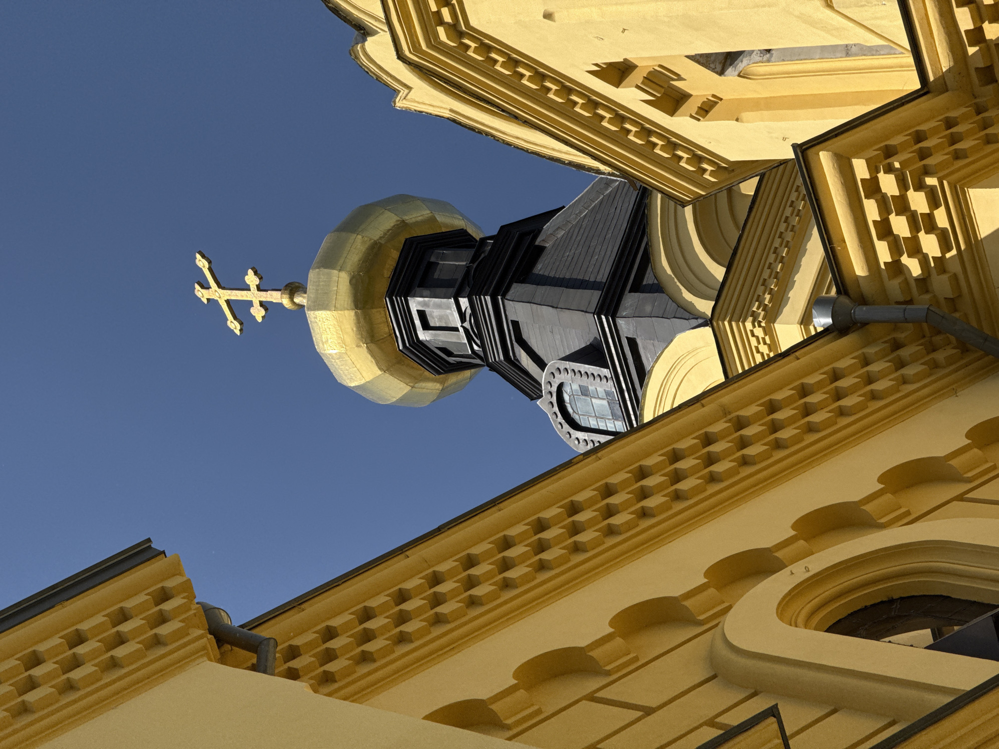 The image is an upward-angled view of a church structure in Nizhny Novgorod, Russia. The architecture features a striking golden dome topped with a cross against a clear blue sky. The building exhibits a warm yellow hue, with intricate geometric patterns and details along the cornices and walls. A drainage pipe is visible on one of the building edges. The dark tower contrasts sharply with the gold and yellow, adding a depth to the scene. A circular window is visible on the tower. The viewpoint emphasizes the towering height and ornate design of the religious structure.