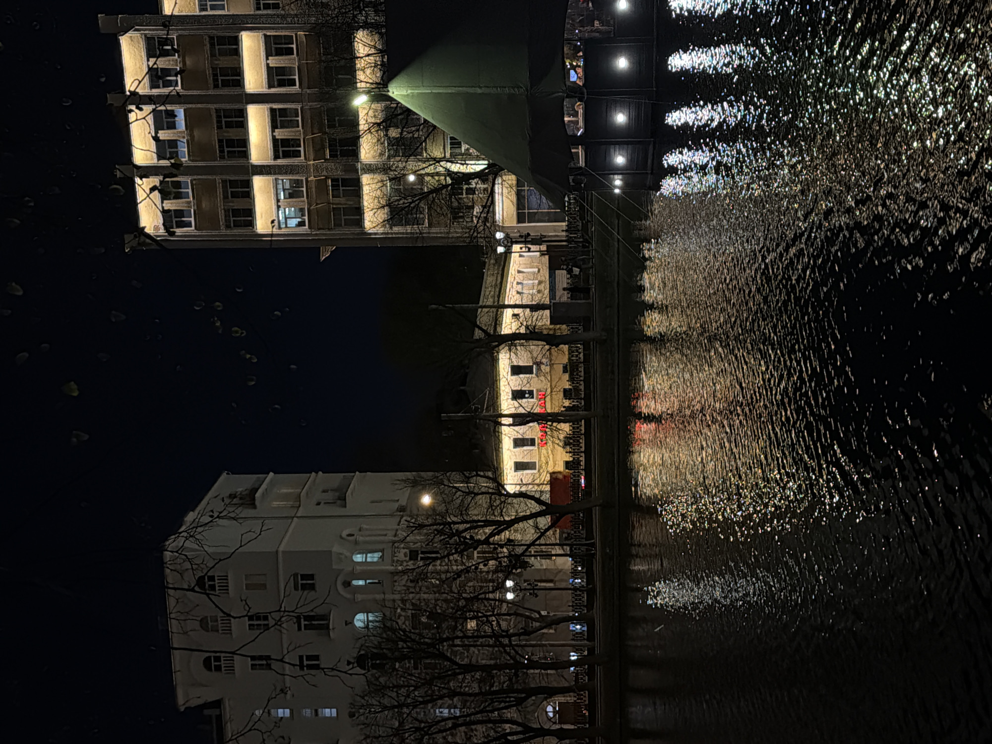 The image shows a nighttime view of the Moscow Canal in Moscow, Russia. A multistory building with lit windows is visible on the left. A bridge with overhead lights and people is in the center. The water's surface reflects light from the buildings and bridge. Bare trees are silhouetted against the buildings.