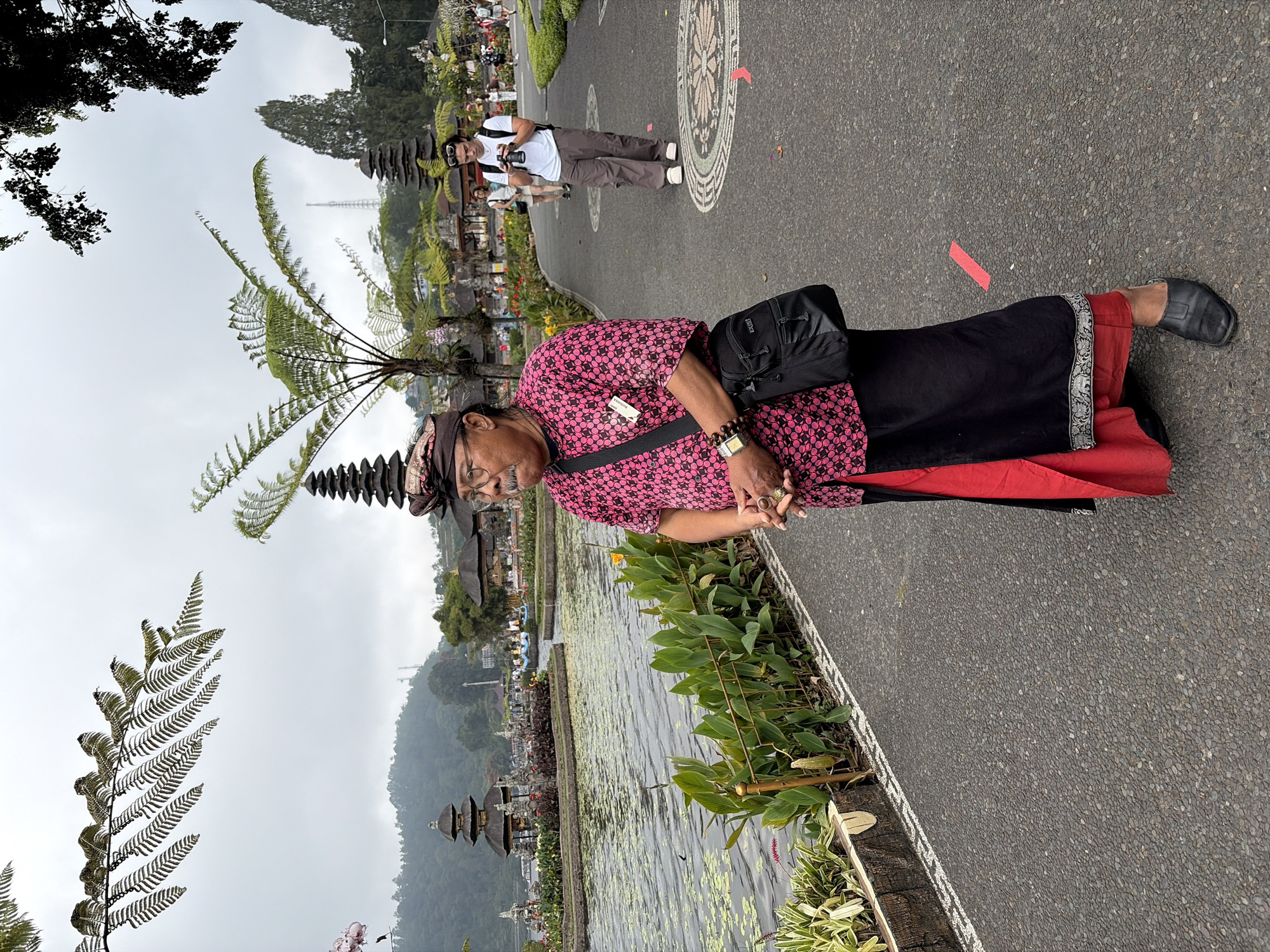 In the city of Baturiti, Indonesia, at the Pura Ulun Danu Batur temple complex on the shores of Lake Batur, an older man with dark skin and a mustache, wearing a patterned pink shirt, black sarong with a red underskirt, a patterned headdress, and black shoes, stands facing left and looking forward with his hands clasped. He has a black crossbody bag over his right shoulder. In the background, across a paved walkway and lush greenery, a large lake is visible, dotted with lily pads and reflecting the overcast sky. Several traditional Balinese temples with tiered roofs are situated along the water's edge and on the surrounding hills. Other people, including a young man holding a camera, are visible walking on the paved path. The environment is lush with various plants, including ferns and flowers, and the atmosphere appears peaceful.
