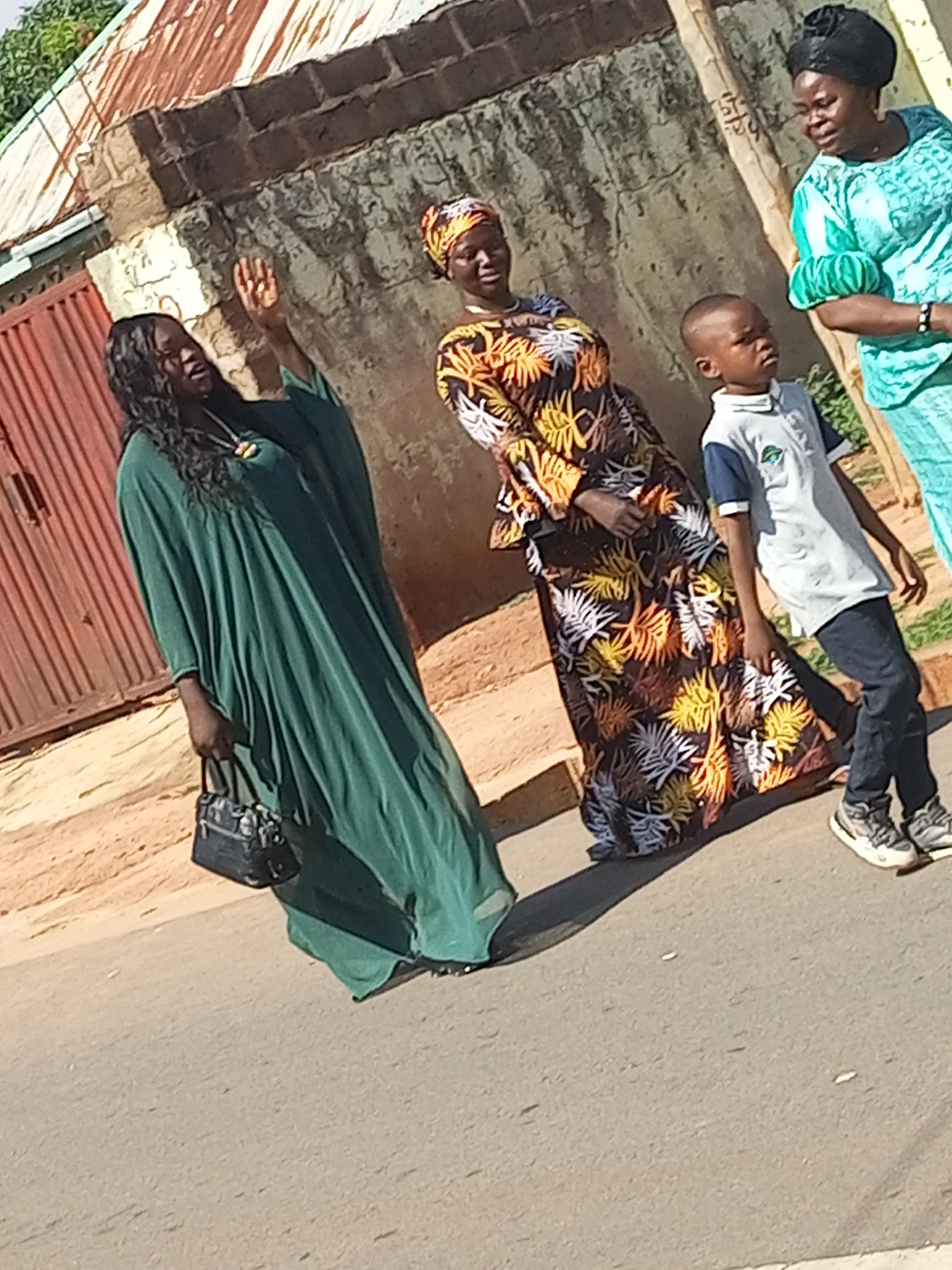 The image depicts an outdoor scene in an urban environment. In the foreground, a woman wearing a long, flowing green dress and holding a black handbag walks towards the left of the frame. Her right hand is raised, and she appears to be speaking or gesturing. Behind her, a woman in a patterned dress with yellow, orange, and white floral motifs, and a headscarf, stands facing the right. To the right of the woman in the patterned dress, a young boy in a gray polo shirt and dark pants walks towards the right. Further to the right, a woman in a turquoise-colored outfit and headscarf is partially visible, facing the left. The background shows a building with a corrugated metal roof and a brick wall. The ground is paved and appears to be a street or sidewalk. The overall lighting suggests it is daytime.