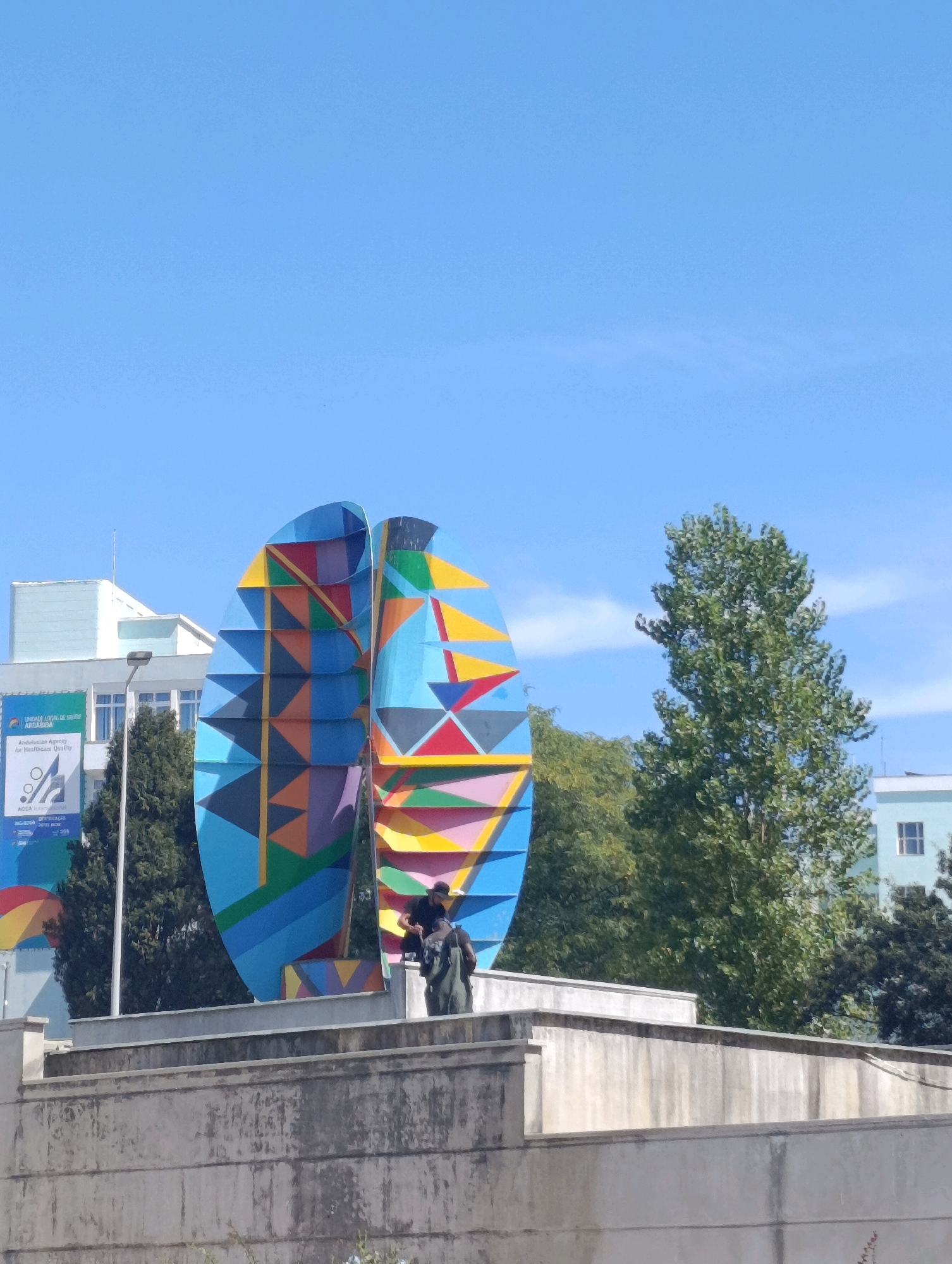 This image captures a section of a large, colorful, abstract sculpture located outdoors in Setúbal, Portugal. The sculpture, which is predominantly blue with geometric shapes in various vibrant colors like yellow, red, orange, and purple, appears to be composed of curved panels. Two individuals are visible on top of a concrete structure in front of the sculpture; one, wearing a dark cap and backpack, is interacting with the sculpture, while the other, also wearing a backpack, stands nearby. Behind the sculpture, a tall, leafy green tree stands prominently against a clear blue sky, with another, less distinct tree to its left. In the background, a modern building with a light blue facade and windows is partially visible, as is a white building with a blue banner. The scene is set on a bright, sunny day, with a clear sky and the hint of a few wispy clouds.