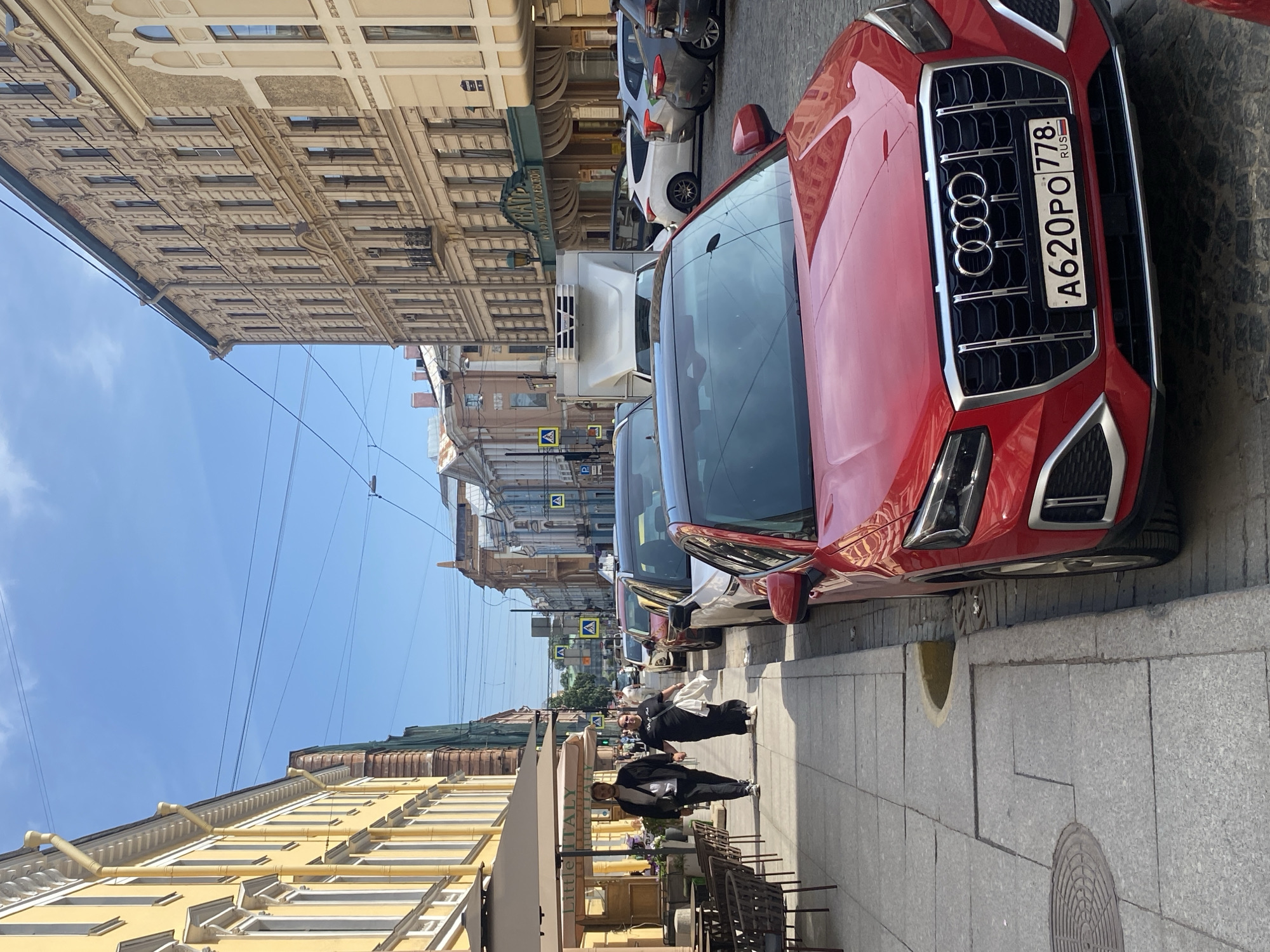 A wide, eye-level shot captures a sunlit street in Saint Petersburg, Russia, with a red Audi Q2 parked in the foreground. The street features traditional St. Petersburg architecture with yellow and beige buildings lining both sides. On the left, outdoor seating for a restaurant, possibly named "Little Italy," is visible beneath large beige umbrellas. A man and a woman walk hand-in-hand on the sidewalk, facing the camera. The street is lined with parked cars, and traffic signals and overhead wires are present. The sky is clear blue with a few wispy clouds. The red Audi's license plate reads "A620PO 778 RUS."
