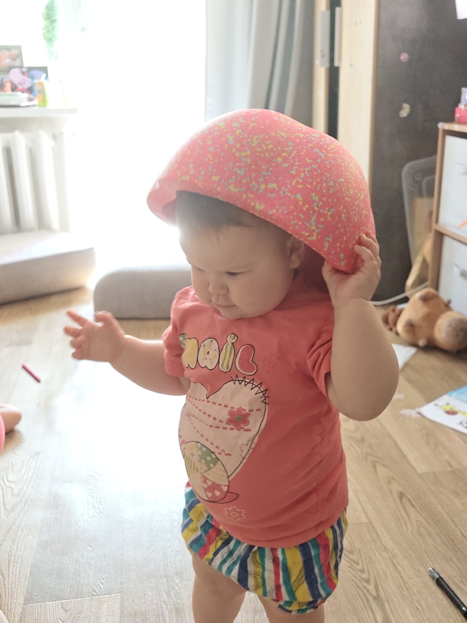 A baby stands on a wooden floor wearing a red, short-sleeved shirt with a cartoon heart design and colorful striped shorts. The baby has a large, red, dome-shaped object on their head and is holding the object with their right hand. The location is indoors, potentially a residence, in the city of Chaikovsky, Russia. Natural light streams in from a window in the background. A radiator and a portion of a gray sofa are visible near the window. Other objects in the room include a plush camel, a child's drawing on the floor, and furniture pieces. The baby appears to be examining something with their left hand extended. A small red crayon is on the floor to the left of the baby.