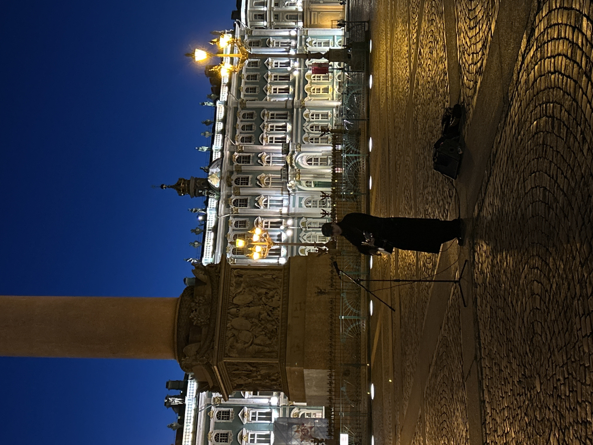 The image depicts a scene in Saint Petersburg, Russia, at night. A street performer in dark clothing is standing on a cobblestone square, playing a guitar and singing into a microphone on a stand. Behind the performer is a large column with decorative carvings, likely part of a monument or architectural feature. In the background, a building with illuminated windows and ornate detailing is visible. The sky is a deep blue, indicating it is nighttime. A speaker is placed on the ground, possibly for amplification. The scene suggests a public performance in a historic urban setting.