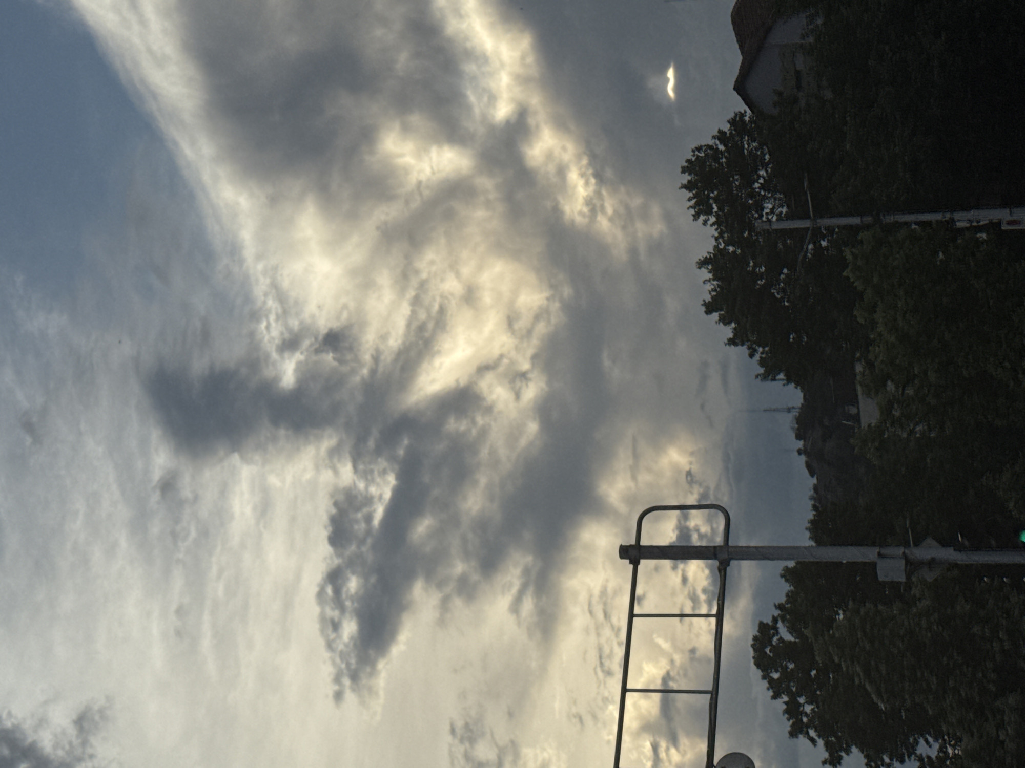This image captures a dramatic sky filled with dark, swirling clouds interspersed with patches of bright sunlight. In the foreground, silhouetted against the sky, are the tops of dark green trees and a portion of a building. A metal pole with a traffic signal or road sign structure is visible on the left side of the frame. The scene appears to be from an urban environment, likely Plovdiv, Bulgaria, given the context of the request. The lighting suggests it could be either early morning or late afternoon, with the sun's rays breaking through the heavy cloud cover. The overall impression is one of an overcast sky with dynamic cloud formations and hints of a coming storm or clearing weather.