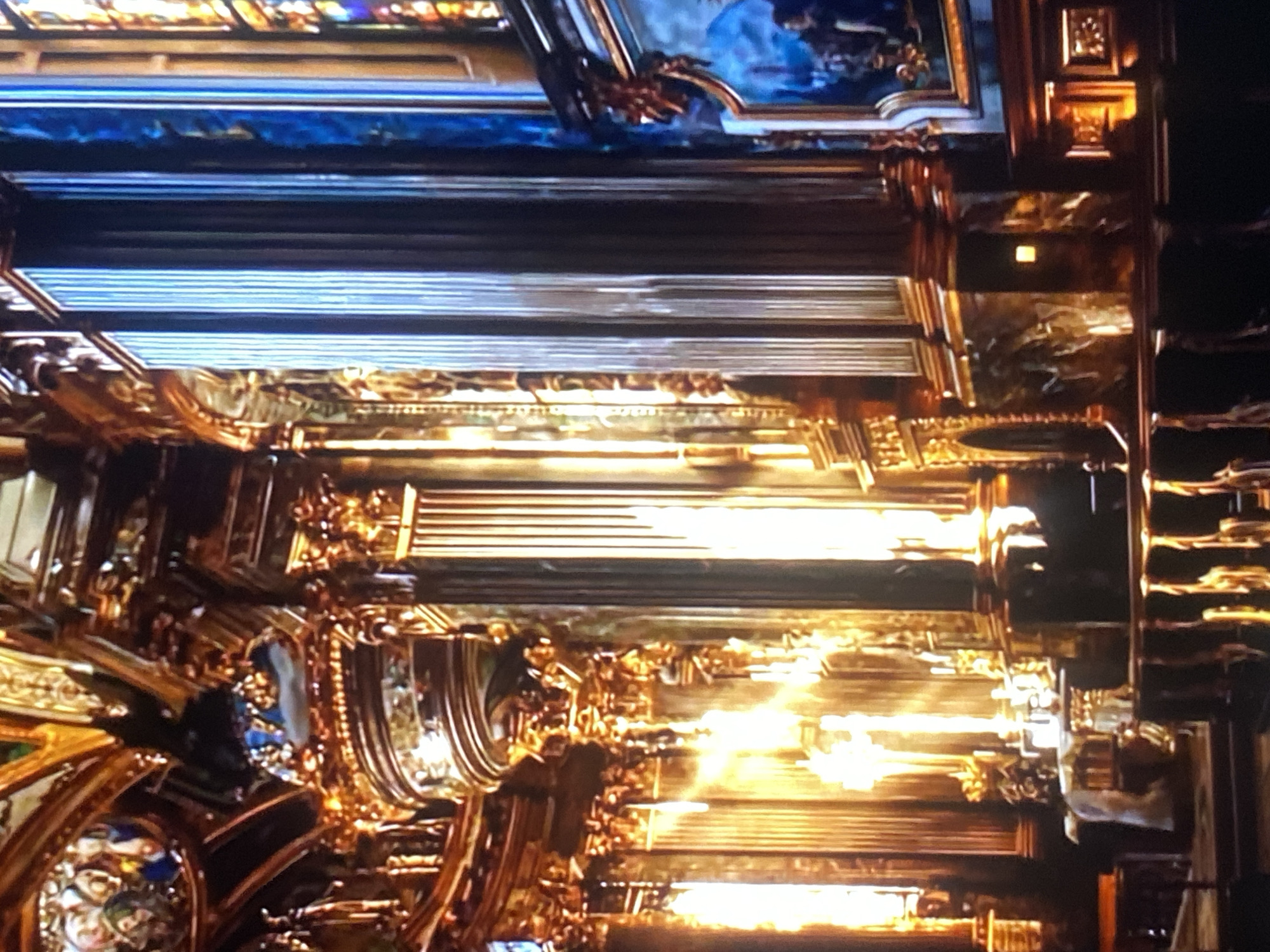 The image shows the interior of a highly ornate building, likely a church or palace, with a focus on the ceiling. The ceiling features intricate gold detailing, paintings, and potentially stained glass. There is strong backlighting, creating a high contrast scene. The image does not provide sufficient information to confirm that the scene is located in Tinaquillo, Venezuela. There are no people or actions visible in the picture.