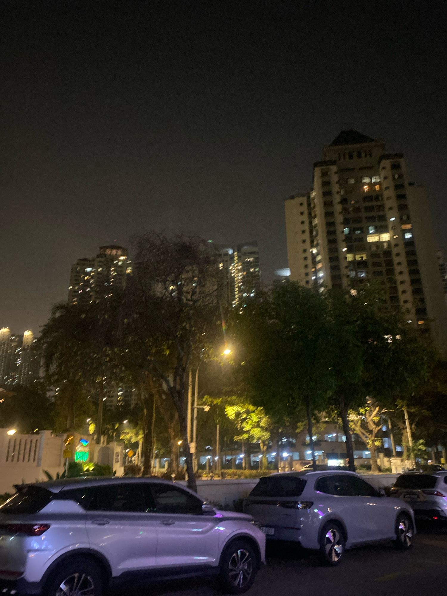 The image depicts a nighttime urban scene in Kuala Lumpur, Malaysia, likely in a residential or commercial district with high-rise buildings. In the foreground, a light-colored SUV, possibly a proton X50 based on its design, is parked. To its right, another similar SUV is partially visible, followed by the rear of a white car, potentially a sedan. The background features several tall apartment buildings with illuminated windows, indicating a populated area. Lush trees and streetlights line the street, adding to the ambient lighting of the scene. The overall atmosphere is one of a quiet, well-lit city evening.