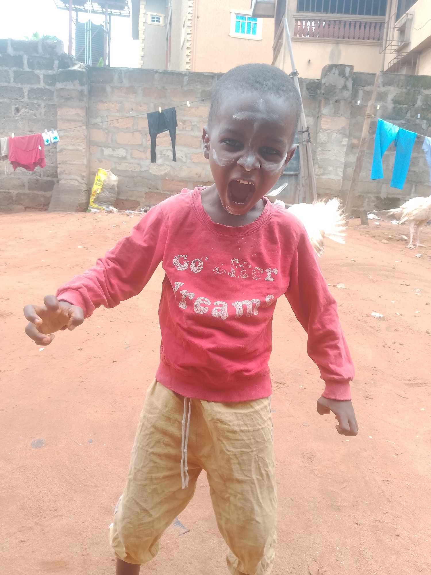 A young child stands in a dirt yard, wearing a red long-sleeved shirt with white lettering and tan pants. The child has white paint or powder on their face and is yelling with their mouth open. A clothesline is visible in the background, with clothes hanging on it. Behind the child is a brick wall and a few buildings. A white chicken stands in the background to the right. The location appears to be outside in an urban area. The city and region is Unknown, Unknown.