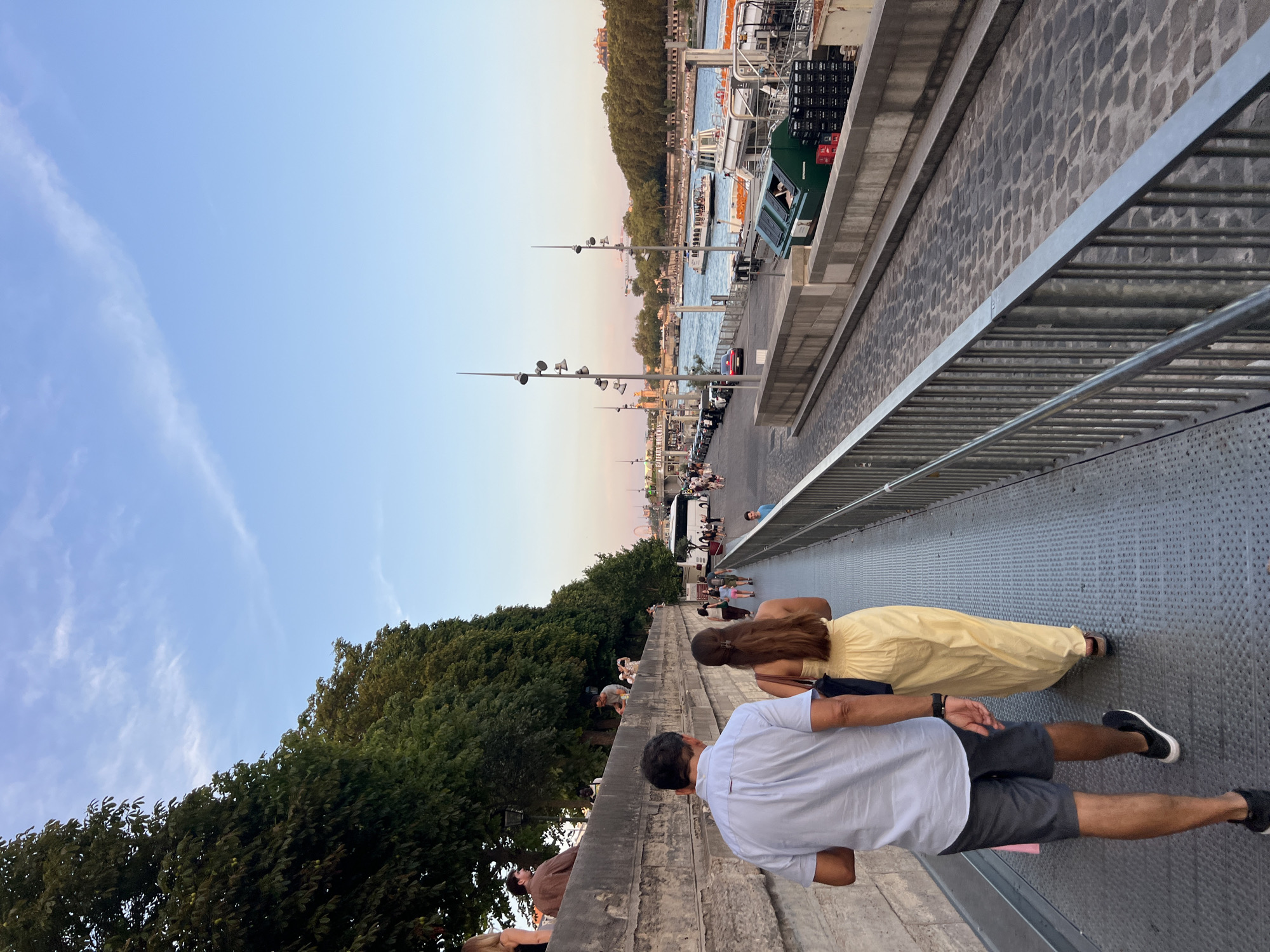 The image captures a view along the Seine River in Paris, France, likely during the late afternoon or early evening, given the soft lighting. In the foreground, a man and a woman walk away from the viewer on a metal walkway. The man is wearing a light blue short-sleeved shirt and dark shorts, while the woman is in a long, flowing yellow dress. They are walking on a raised, grated metal pathway with a metal railing on the right.
Beyond the walkway, a cobblestone path runs parallel to the river. A row of parked cars is visible along this path. The Seine River itself is to the right, with a tour boat carrying passengers moving across it. Further in the distance, the Parisian skyline is visible, with buildings and trees lining the opposite bank of the river. Trees and vegetation also line the left side of the walkway, separating it from a stone embankment where other people are seen sitting and walking. The sky above is a clear blue with wispy white clouds. The overall scene depicts a leisurely stroll along the river in a popular tourist area of Paris.