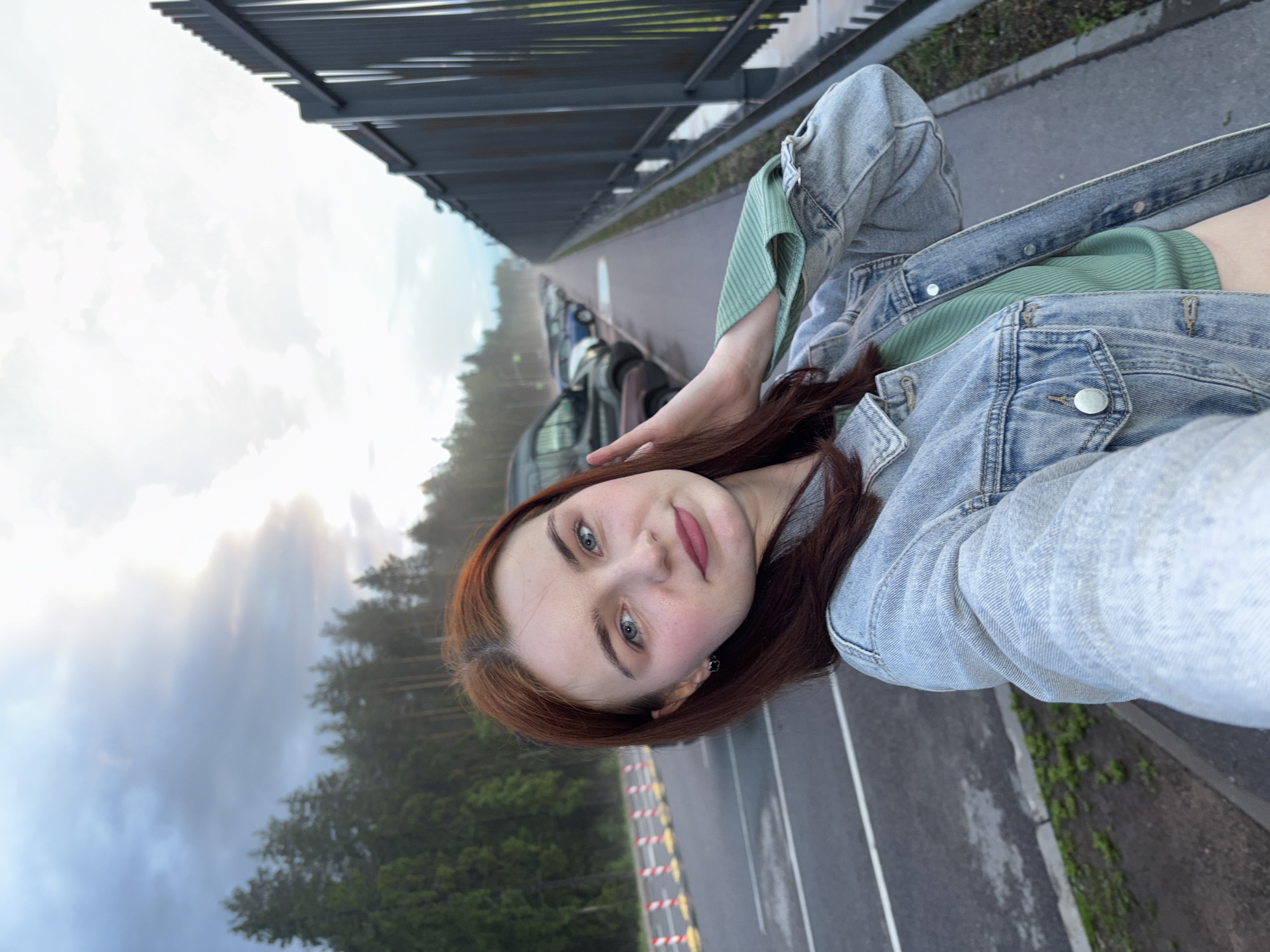 A young Caucasian woman with auburn hair and blue eyes is taking a selfie. She is wearing a light blue denim jacket over a green ribbed top. Her left hand is touching her head. She is standing in an outdoor parking lot with a row of cars parked in the background. To her right, there is a tall, dark metal fence and a sidewalk. To her left, there are trees and a line of orange and white traffic cones. The sky is overcast with some sunlight breaking through. The scene appears to be in the city of Arfa, Russia.