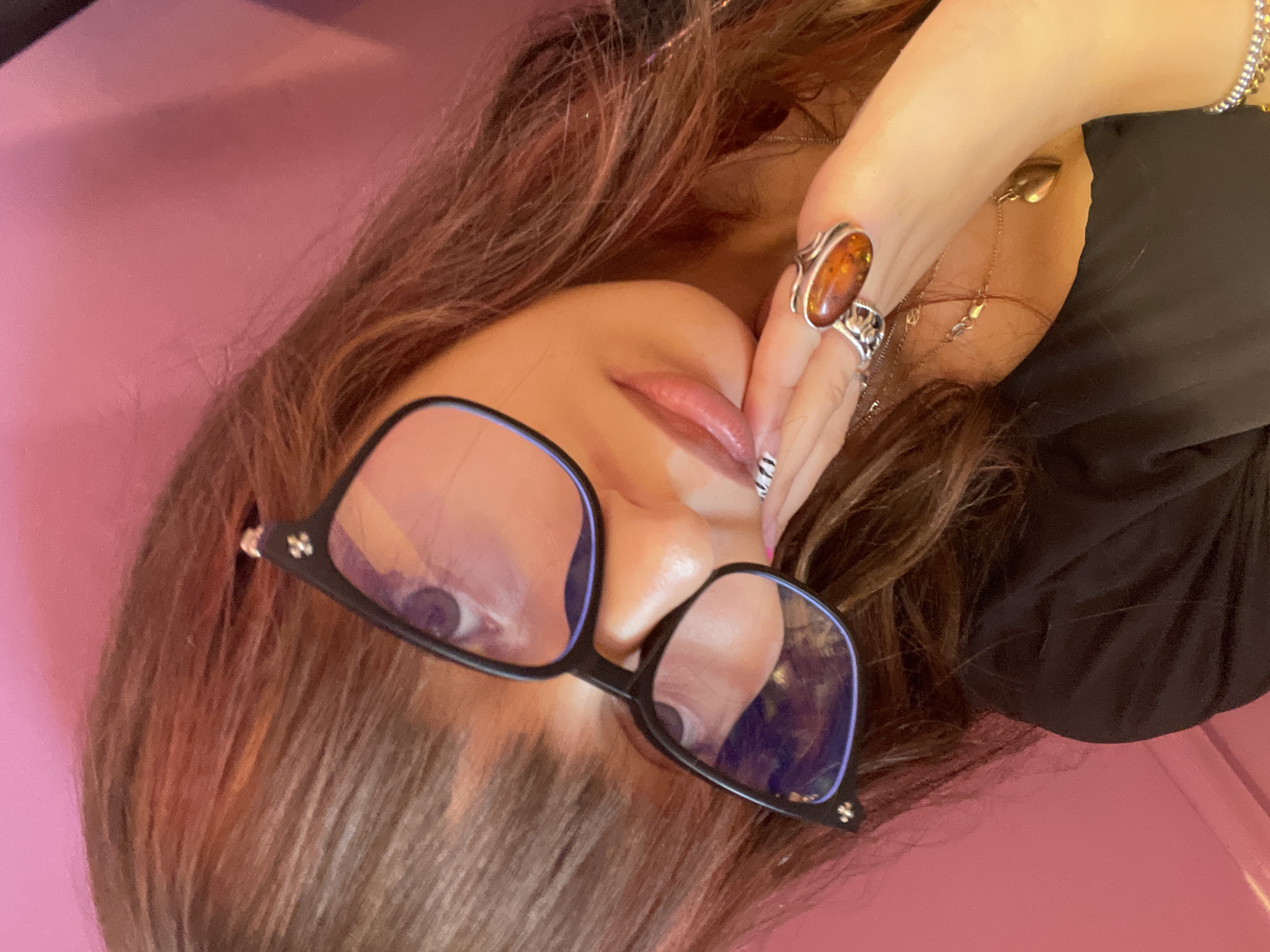 A close-up, high-angle, medium shot captures a person with long, brown hair wearing black-framed glasses and a black top, resting their head on their hand. Their face is partially obscured by the glasses, but their lips are visible, as is a ring on their right index finger. The background is a solid pink color, with some subtle texture. The lighting is soft and warm, casting a gentle glow on the subject. The overall mood is intimate and casual.