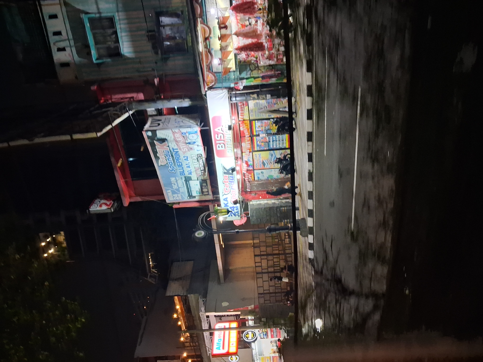 The image depicts a street scene at night in Depok, Indonesia. On the right side of the street, there is a brightly lit store with a red and white awning that displays the text "BISA DITUNGGU". Below this, the store features glass windows showcasing merchandise and multiple red-colored umbrellas hanging outside. To the left of this store, a sign reads "ALFAMART" with its distinctive red and yellow logo. Further to the left, a multi-story building is visible with illuminated windows on the upper floors. In the foreground, the street appears wet and reflects the lights from the shops and buildings, suggesting recent rain. Several motorcycles are parked on the sidewalk in front of the shops. A few individuals are seen standing or sitting near the stores and on the sidewalk. A street lamp with a decorative top is positioned in the middle ground, casting light onto the scene. The overall impression is one of a busy commercial area at night.