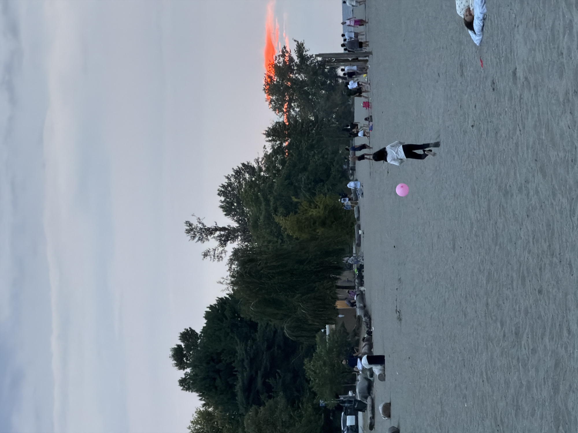 This is an eye-level, medium shot of a beach in Vancouver, Canada, likely Kitsilano Beach due to the presence of trees lining the shore and a general sunset glow. The sand is light brown and textured with small ripples. In the foreground, a young girl with dark hair, wearing a white long-sleeved shirt and black pants, is running barefoot to the left, chasing a pink balloon that is in motion. To the right, a person is lying on a blanket, appearing to be asleep.
In the midground, a group of people are scattered across the beach, some standing and talking, others sitting. There are large rocks and logs along the shoreline, with some people gathered around them. A few beach umbrellas are visible. A building or structure is visible behind some of the people, with activity happening near its entrance.
The background features a dense line of dark green trees, characteristic of the Vancouver coastline. Above the trees, the sky is a pale grey, transitioning to a soft orange and pink glow on the horizon, indicating a sunset. The overall atmosphere is relaxed and recreational, with people enjoying the beach as the day winds down.