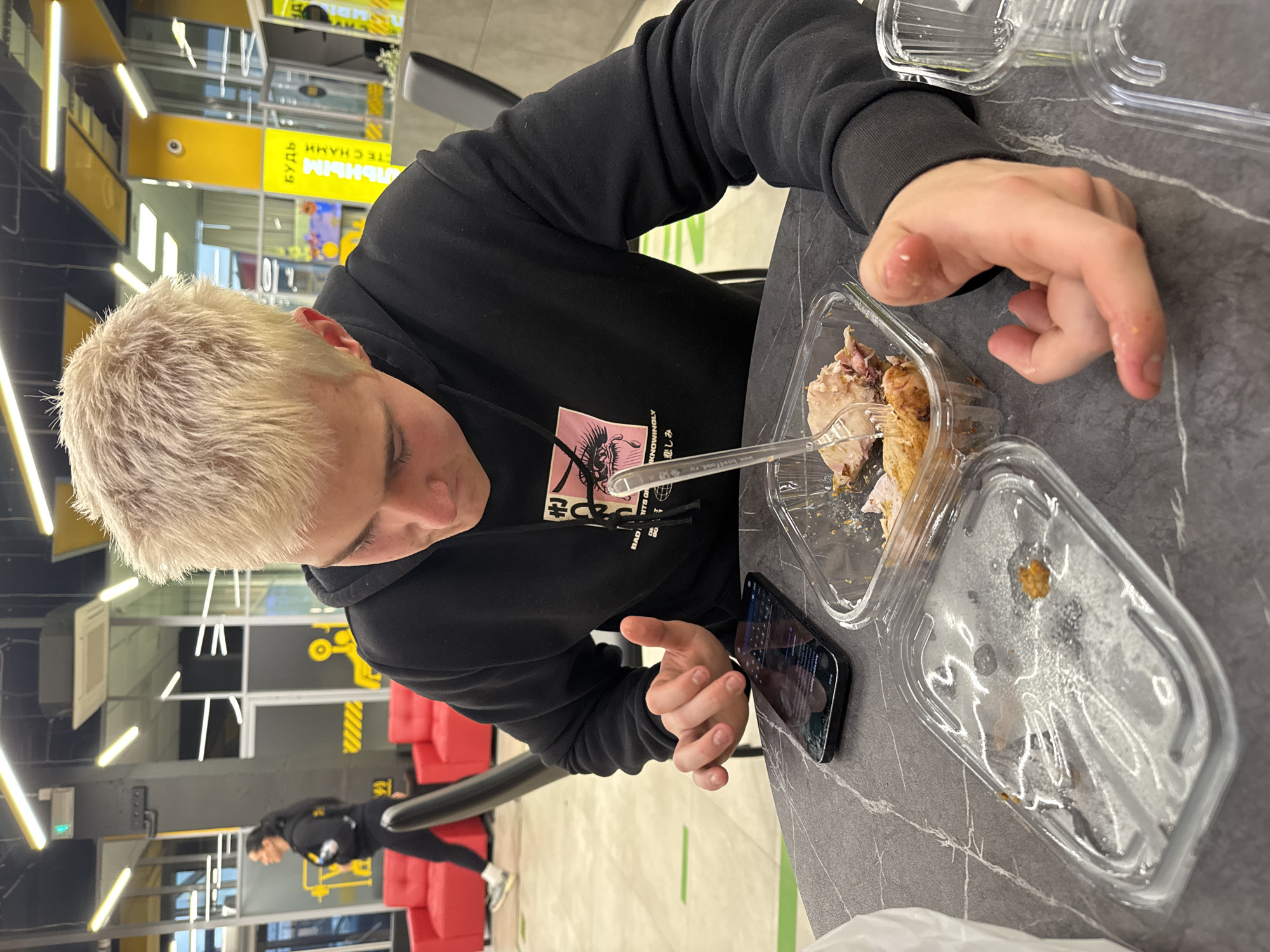 The media file depicts a young person with short, bleached blonde hair and light eyebrows, seated at a dark grey table in an indoor setting. They are wearing a black hoodie that features a graphic design including the English word "KNOWINGLY," the partial text "BAD ITS OR," and the Japanese Kanji for "sadness" or "sorrow" (悲しみ).
The main subject is actively eating from a clear plastic container filled with pieces of cooked chicken or meat, using a clear plastic fork. An empty, clear plastic lid from the container rests beside their meal. A smartphone lies on the table near their left hand. The food container has "box4food.ru" printed on it.
The background suggests a modern, commercial interior, possibly a food court, cafeteria, or a lounge area within a larger facility like a gym. Dark walls are accented with bright yellow panels, one of which displays upside-down Cyrillic text that reads "БУДЬ АКТИВНЫМ С НАМИ" (Be active with us), indicating a Russian-speaking location and hinting at a fitness-oriented environment. Fluorescent strip lights illuminate the space. Red couches are visible in the mid-ground, and another individual, dressed in black, can be seen standing or walking further back. Green tape lines are present on the light-colored floor. The primary subject appears focused on their meal and phone.
