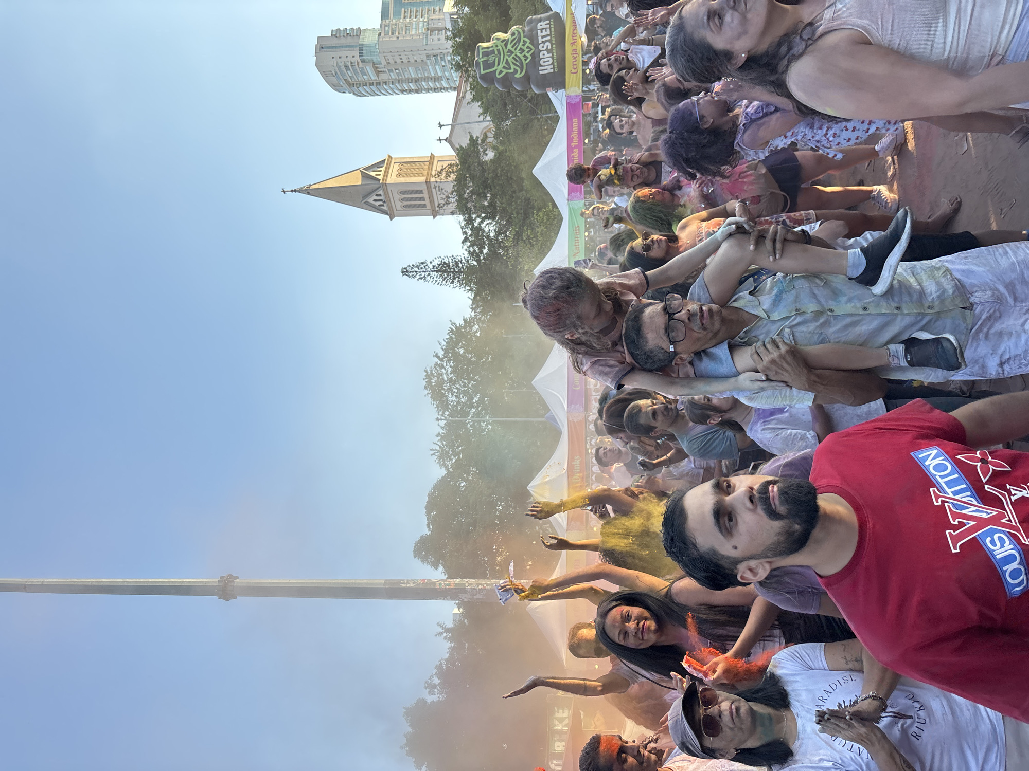 The image captures a crowded scene in a public outdoor space, likely during a Holi festival celebration. A multitude of people are present, their faces and clothes covered in colorful powder, indicative of the event. In the foreground, several individuals are clearly visible, one wearing a red t-shirt with a prominent designer logo. In the background, there is a church steeple visible, along with some modern buildings. The festival is happening in the city of São Paulo, Brazil. Banners and signage are scattered throughout the scene, including "HOPSTER CERVEJARIA." The overall atmosphere is festive and energetic.
